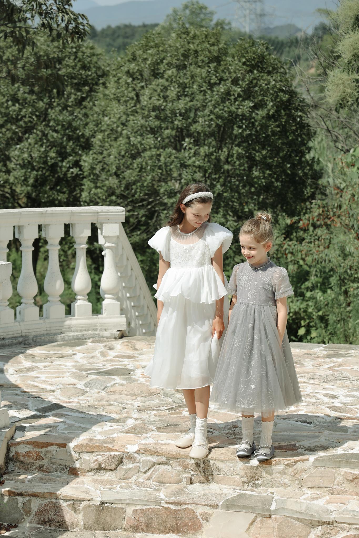 Two young girls in formal dresses walk down stone steps outdoors, one wearing the KIKI and JOJO Girls Diamanté Padded Hairband in Ivory, surrounded by greenery and a white balustrade.