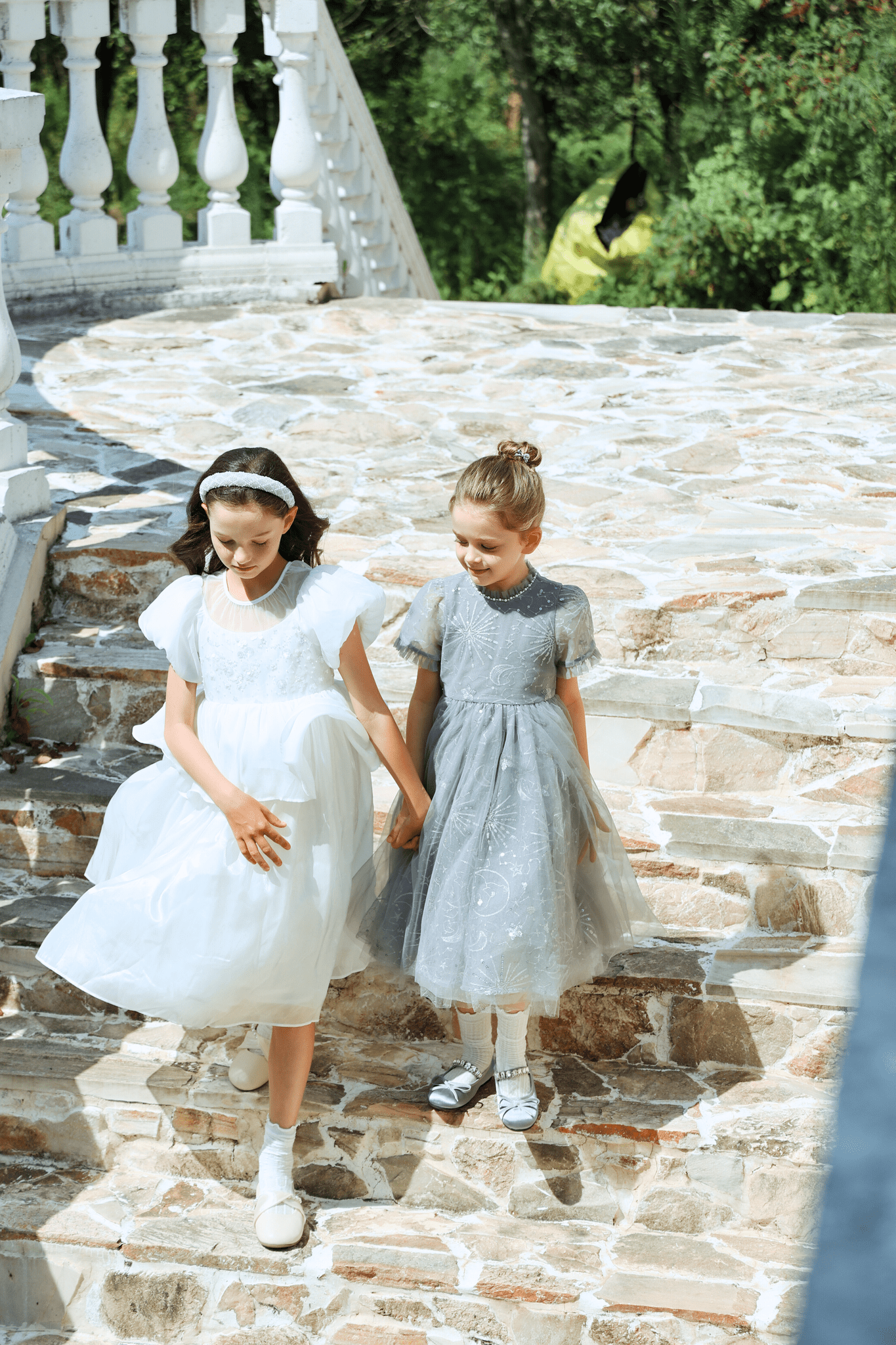 Two young girls in formal dresses wear KIKI and JOJO Girls Plated Metal Bow Hair Clips (2 Pack) - Silver as they walk hand-in-hand down stone steps outside, with greenery and a white railing behind them.