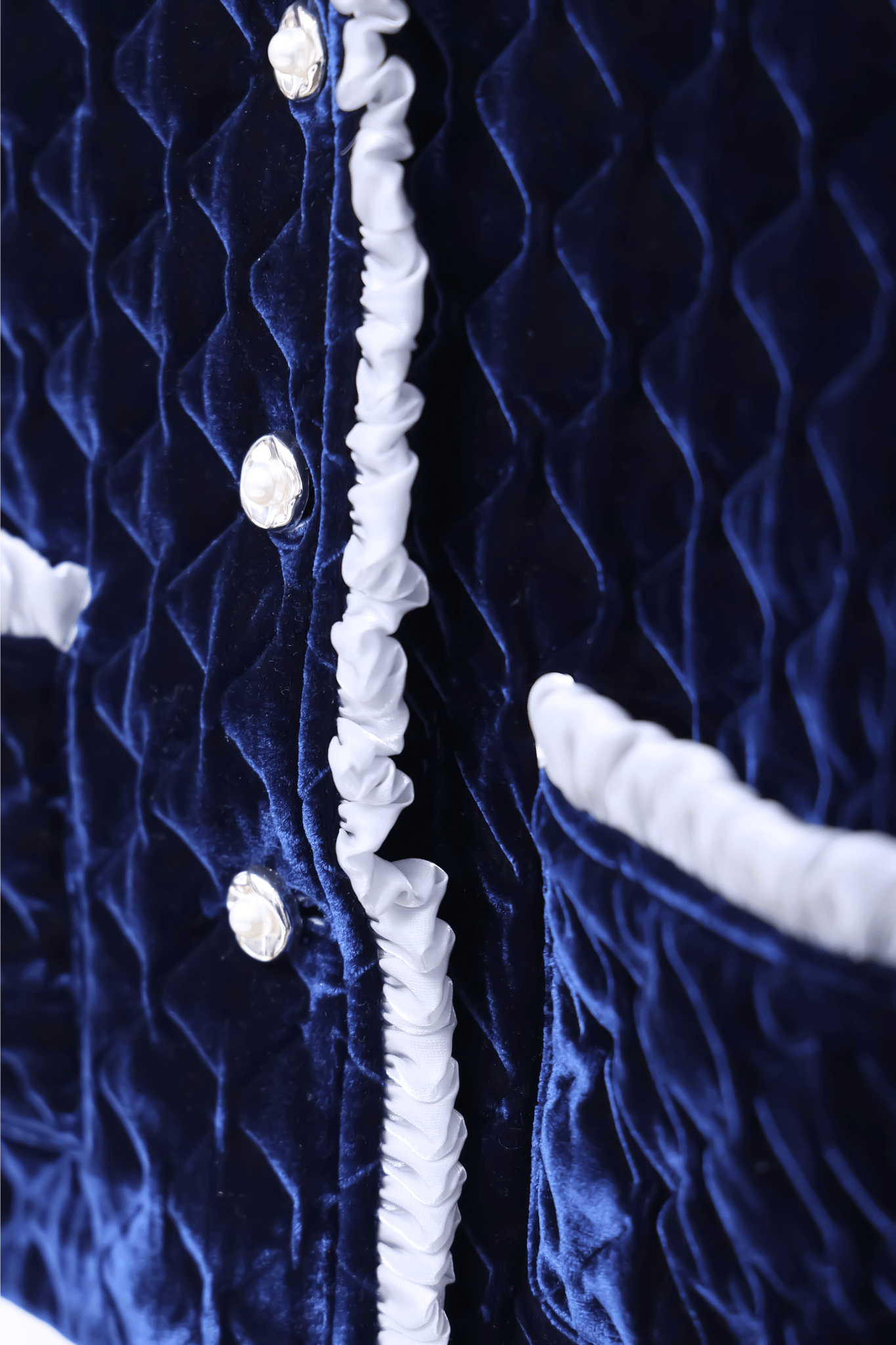 The Girls Quilted Ruffle Jacket in Sapphire Blue by KIKI and JOJO showcases diamond-quilted velvet, petal-shaped buttons, white ruffled trim, and two front pockets.