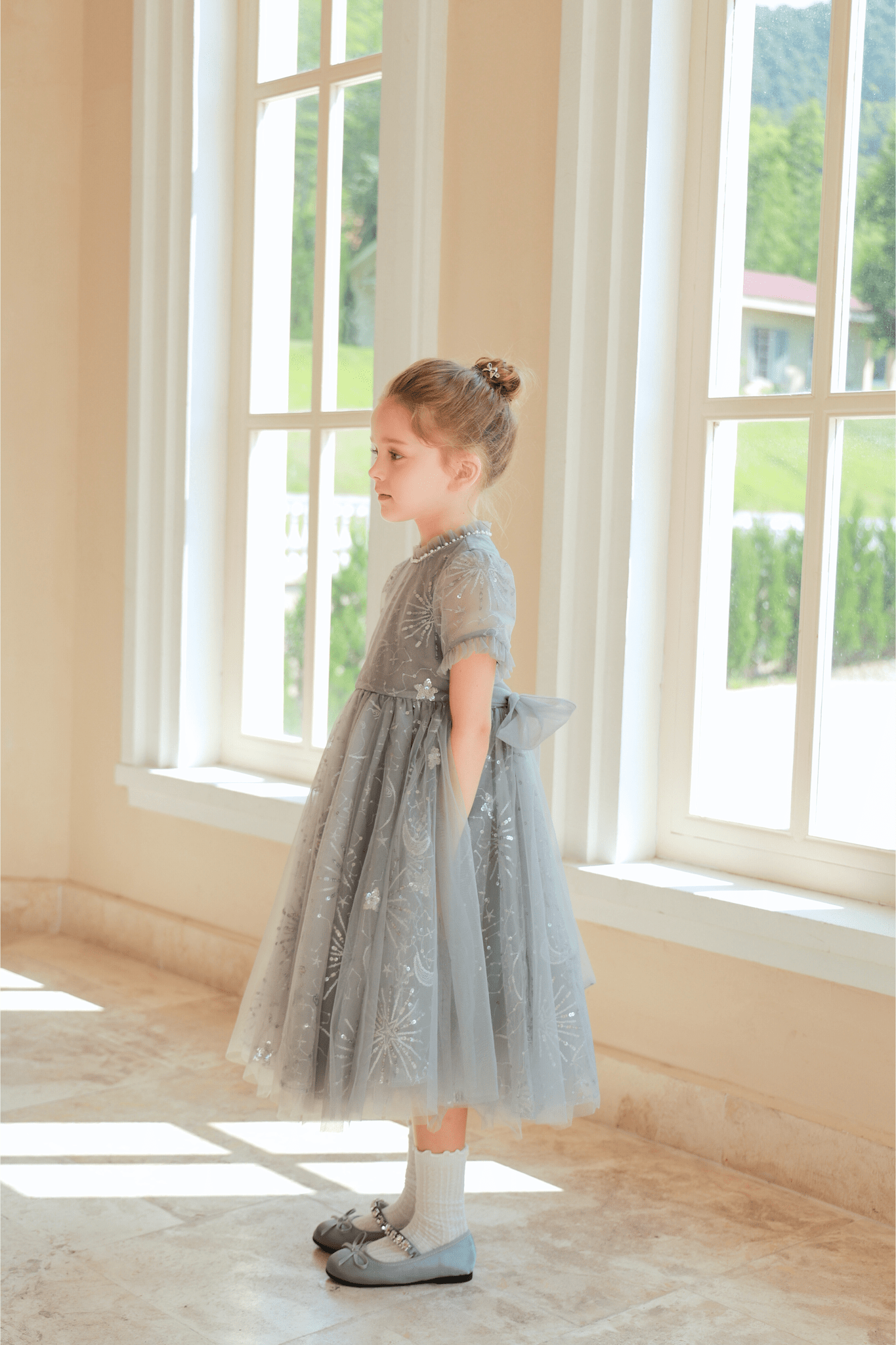 A young girl in a KIKI and JOJO Girls Sequin Embroidered Tulle Dress - Starlight Silver stands sideways by large windows in a bright room, gazing outside.