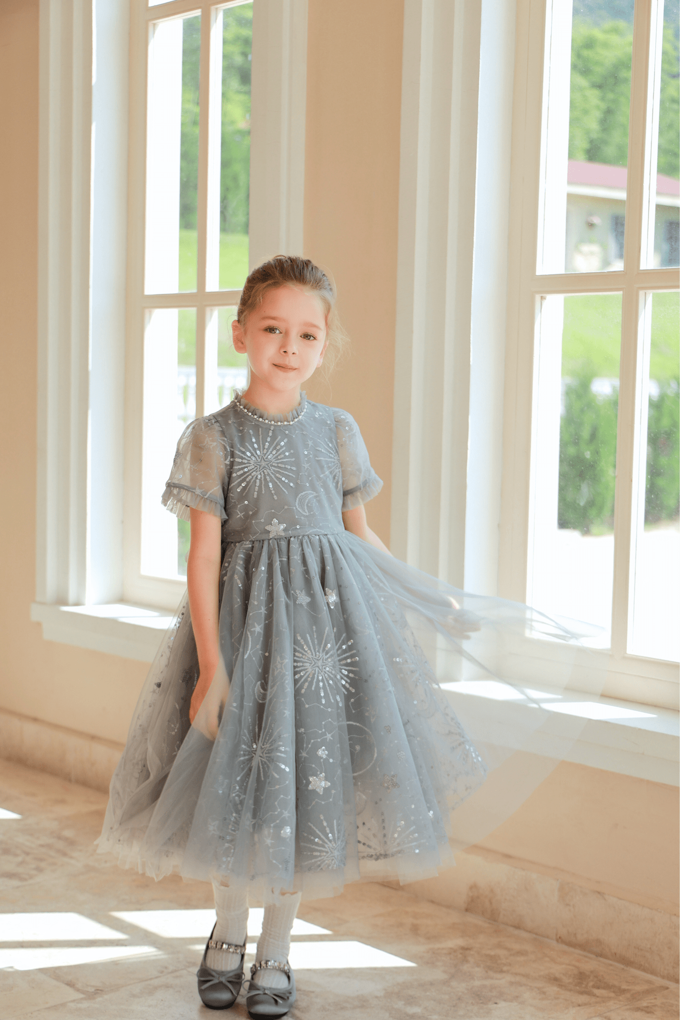 A young girl stands indoors by large windows, lightly holding her sparkly Girls Sequin Embroidered Tulle Dress - Starlight Silver from KIKI and JOJO.