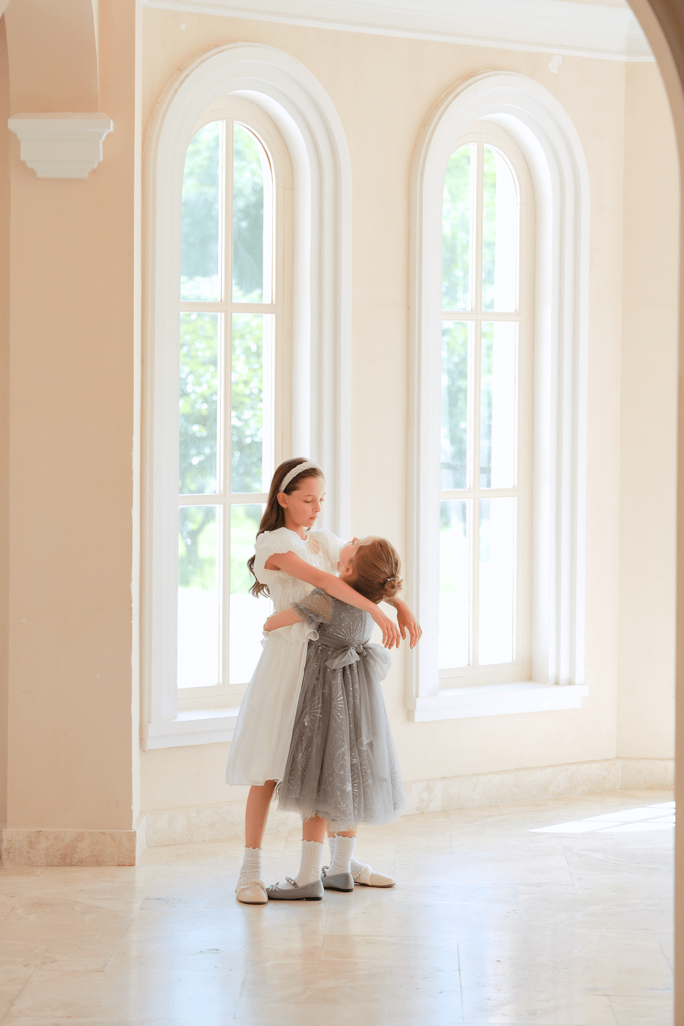 Two young girls, one wearing the KIKI and JOJO Girls Sequin Embroidered Tulle Dress in Starlight Silver, stand embracing before tall arched windows in a bright, sunlit room.