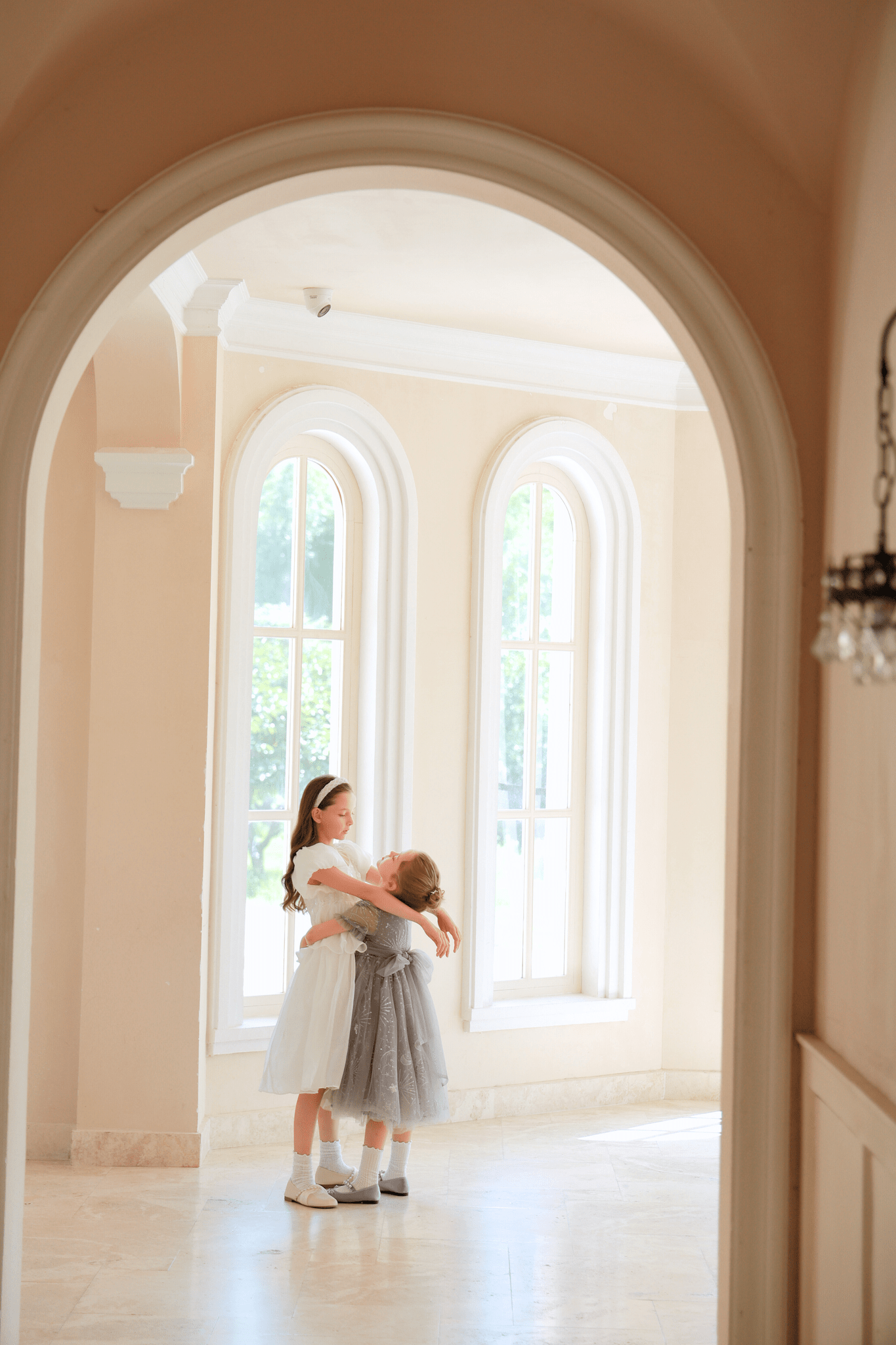 Two young girls, one wearing the KIKI and JOJO Girls Sequin Embroidered Tulle Dress in Starlight Silver, hug each other in a sunlit room with tall arched windows and cream-colored walls.