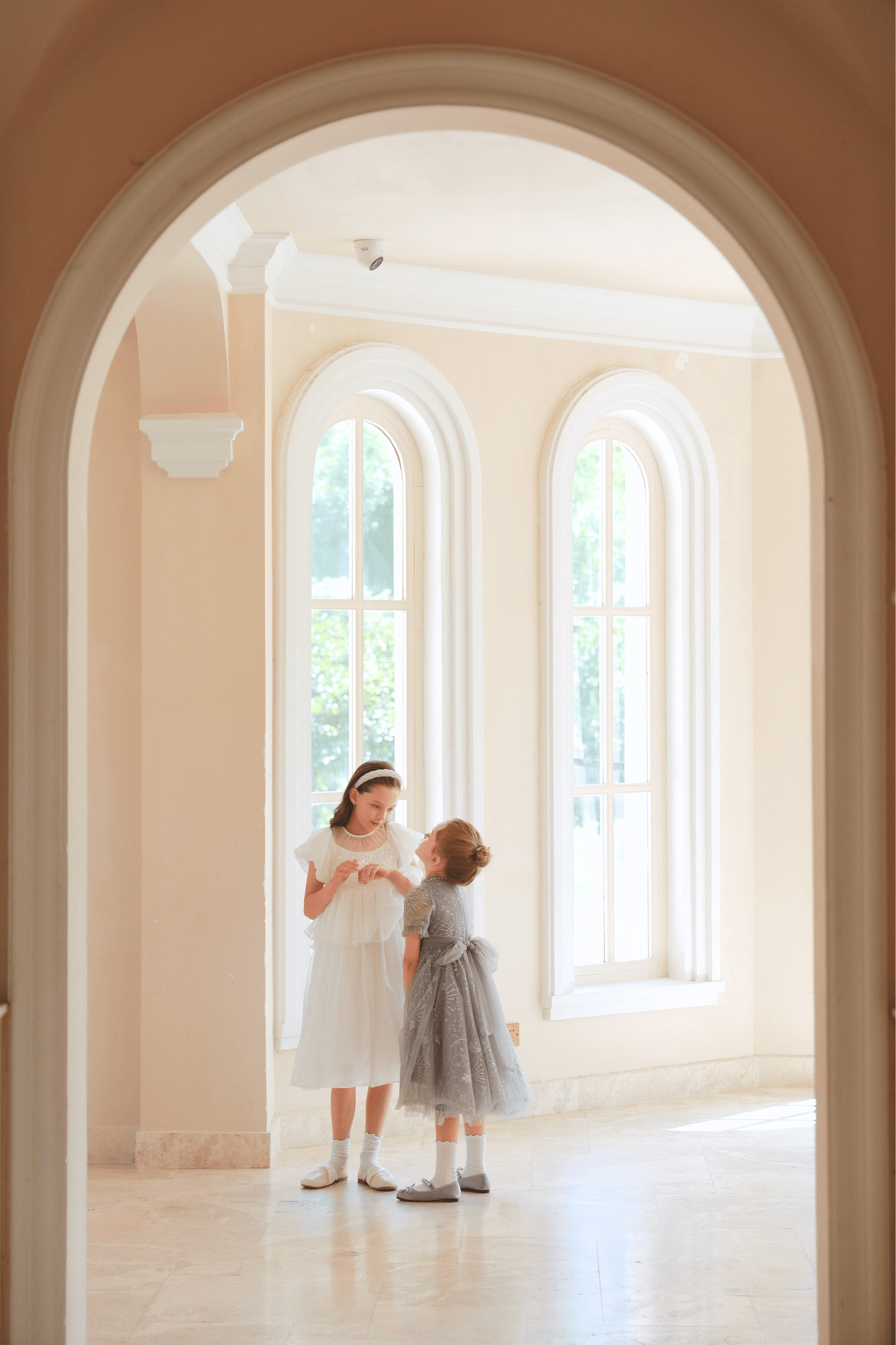 Two young girls stand and talk in a bright, arched room. One wears the KIKI and JOJO Girls Sequin Embroidered Tulle Dress in Starlight Silver, featuring sparkling sequins, as sunlight pours through tall windows onto marble floors.