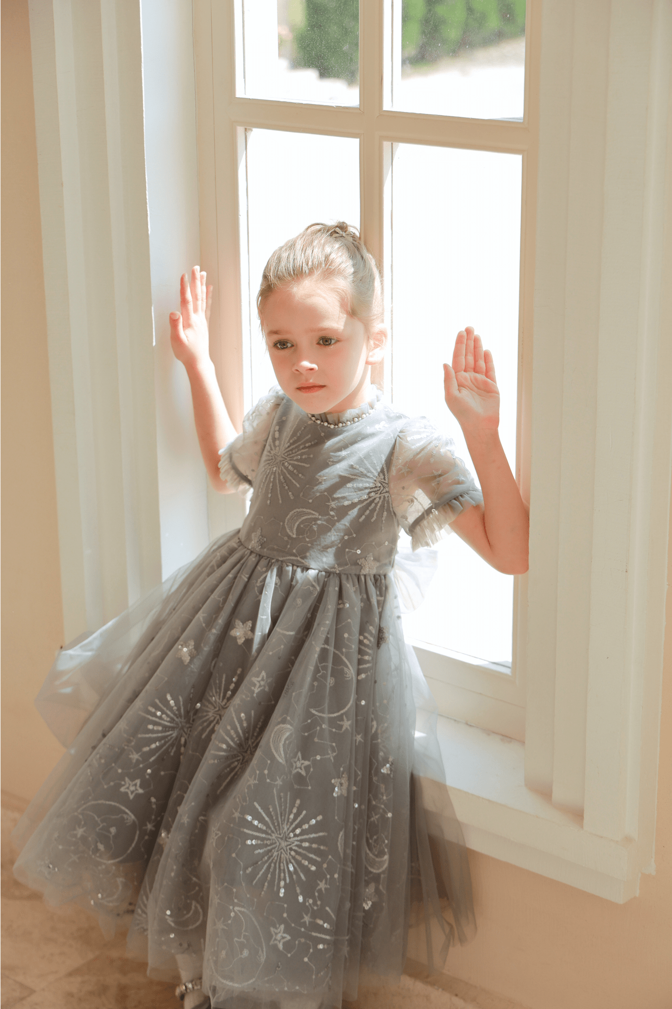 A young girl in the KIKI and JOJO Girls Sequin Embroidered Tulle Dress – Starlight Silver stands by a sunlit window, her hands resting on the frame.