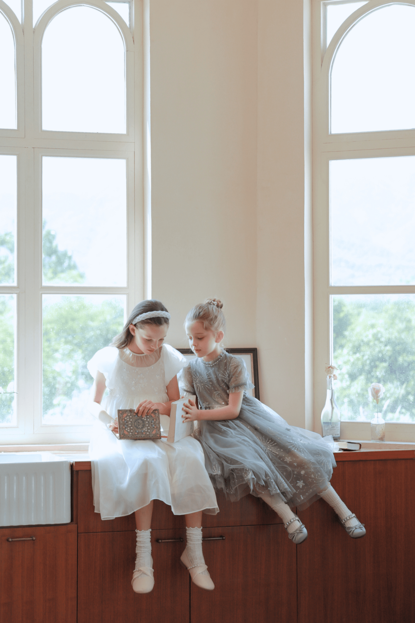 Two young girls in white and a Girls Sequin Embroidered Tulle Dress - Starlight Silver by KIKI and JOJO sit by tall windows on a wooden cabinet, looking together at a sparkling book with sequin and hand-embroidered details.