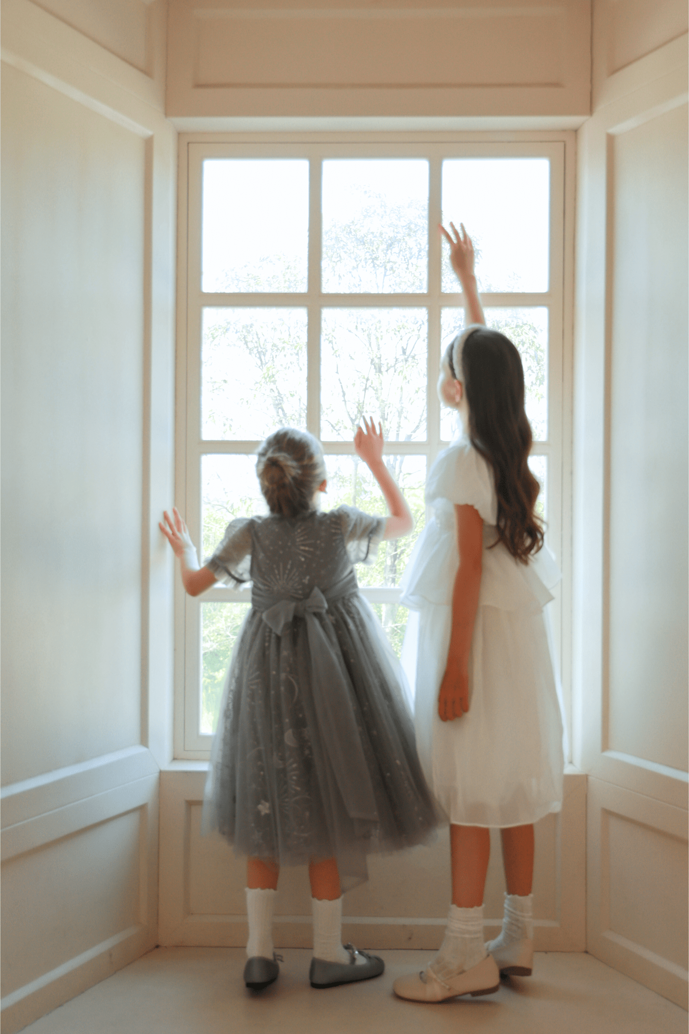 Two young girls stand indoors by a large window, hands on the glass. One wears the KIKI and JOJO Girls Sequin Embroidered Tulle Dress in Starlight Silver, its detailed embroidery shimmering in the light.