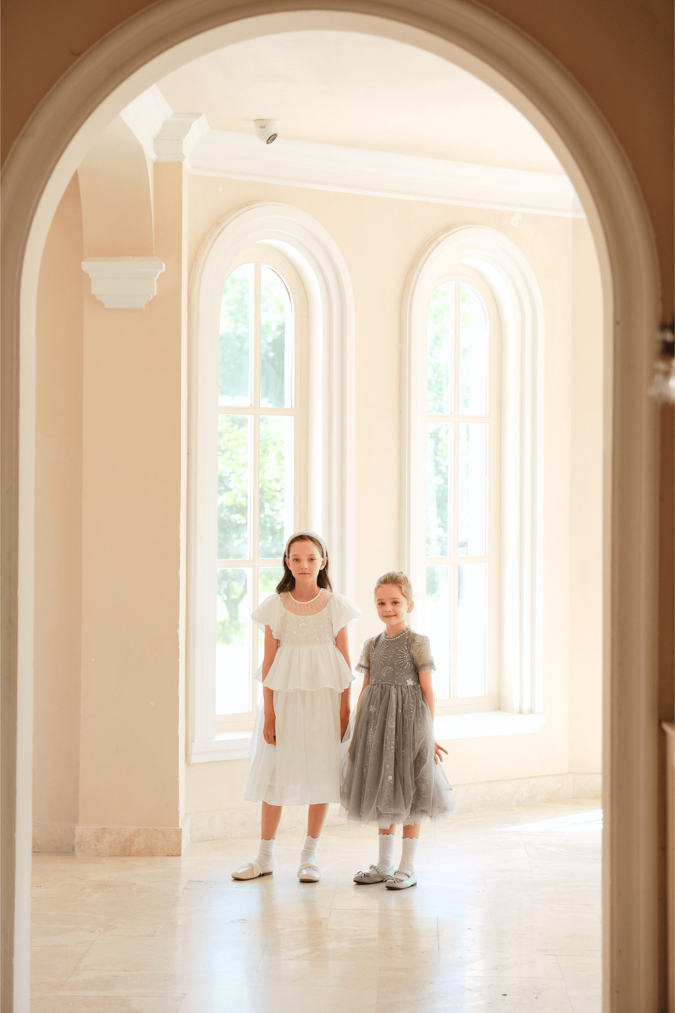 Two young girls stand together in a bright room with large arched windows—one wears white, the other shines in the KIKI and JOJO Girls Sequin Embroidered Tulle Dress in Starlight Silver.