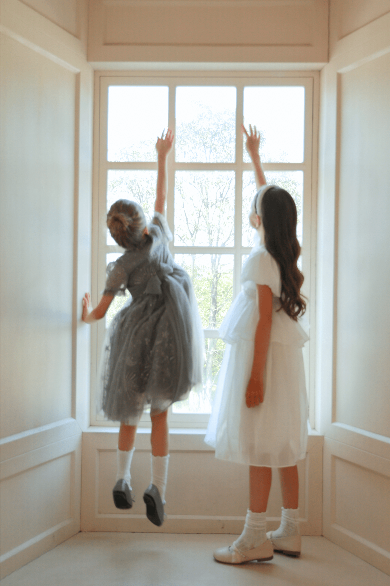 Two girls in dresses stand by a large window; one wears the KIKI and JOJO Girls Sequin Embroidered Tulle Dress in Starlight Silver and jumps, reaching upward, while the other stands beside her as both reach toward the window.