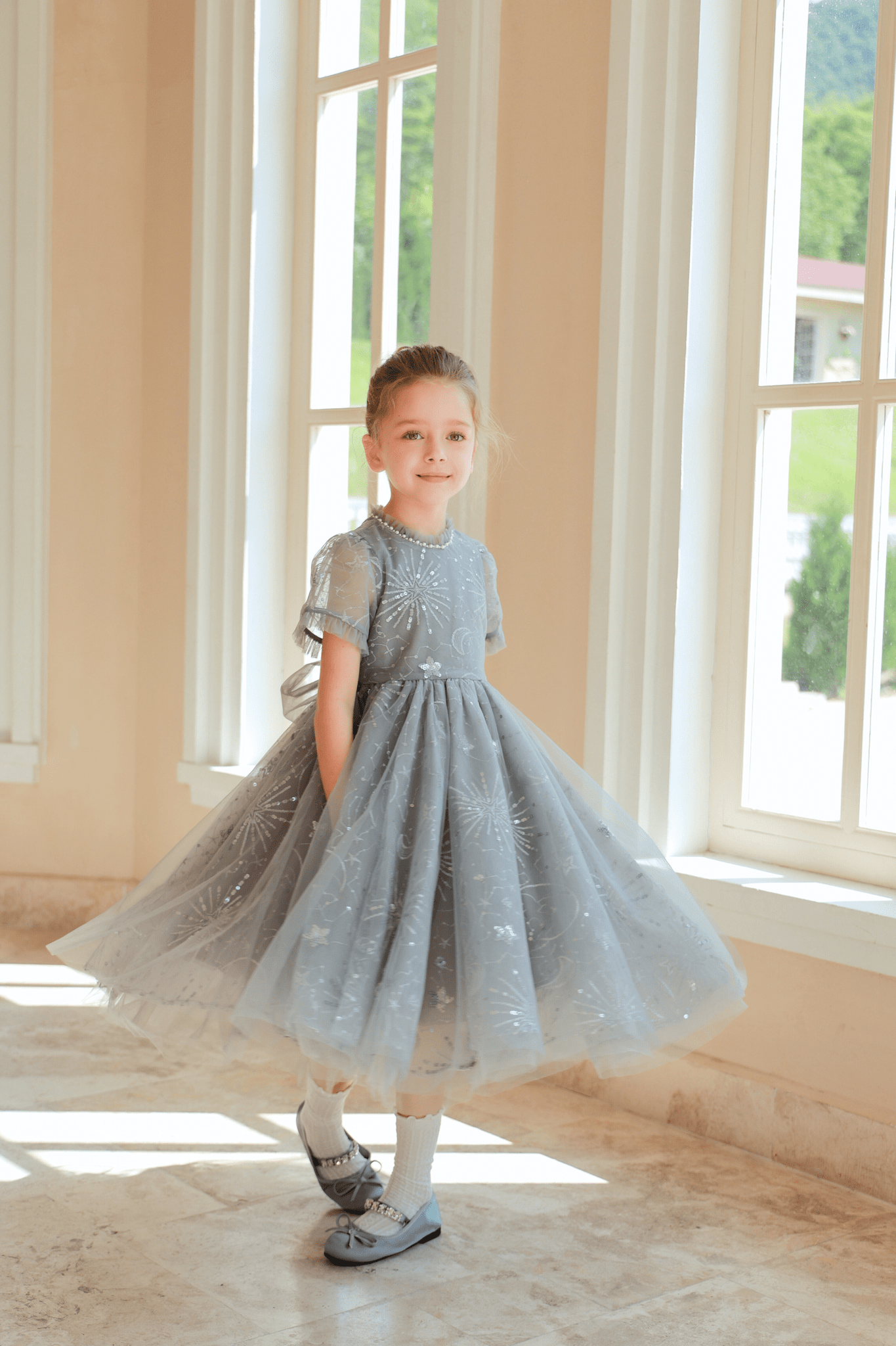 Indoors by large windows, a young girl wears the KIKI and JOJO Girls Sequin Embroidered Tulle Dress in Starlight Silver, featuring hand-embroidered sequins and short sleeves, styled with white socks and blue shoes.