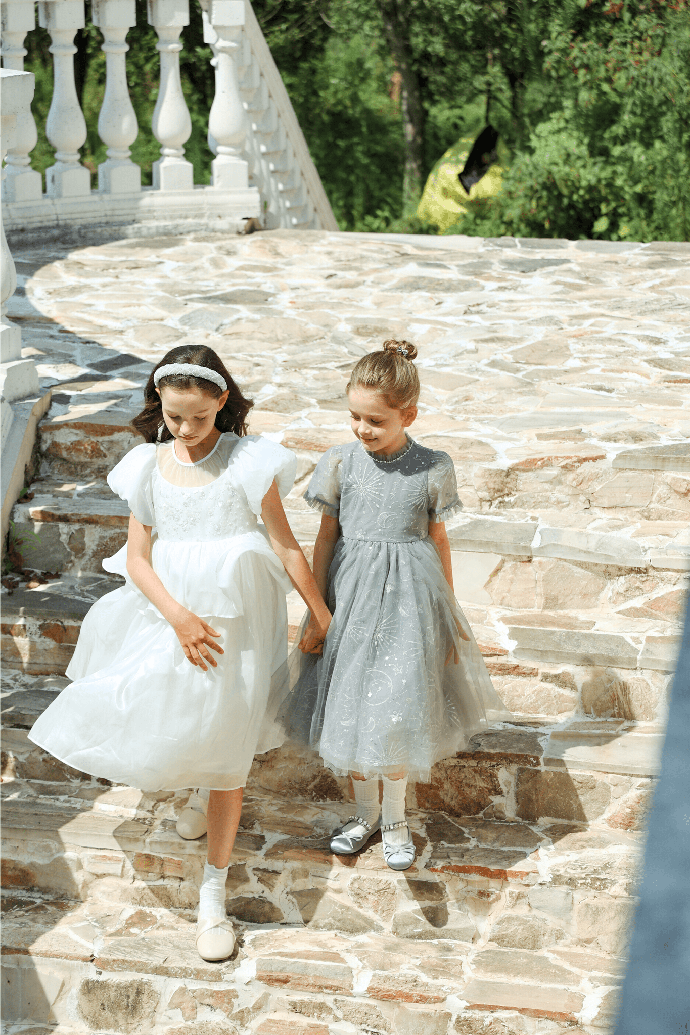 Two young girls walk down outdoor stone steps holding hands. One wears the KIKI and JOJO Girls Sequin Embroidered Tulle Dress in Starlight Silver. Lush greenery and a white railing create a charming backdrop.