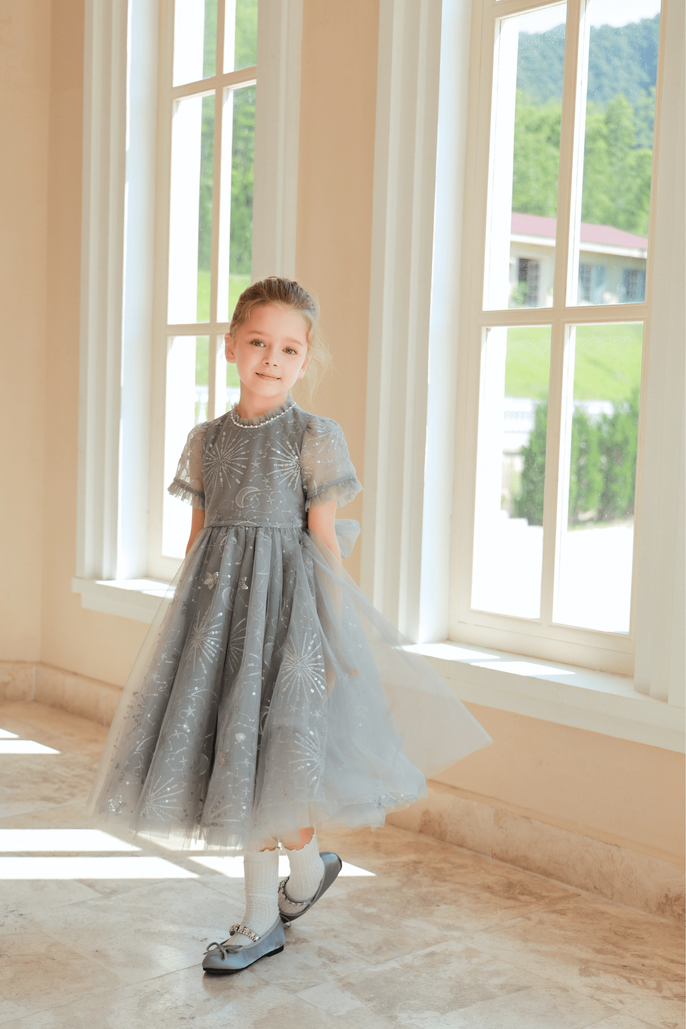 A young girl wearing the KIKI and JOJO Girls Sequin Embroidered Tulle Dress in Starlight Silver stands by large sunlit windows, looking at the camera.