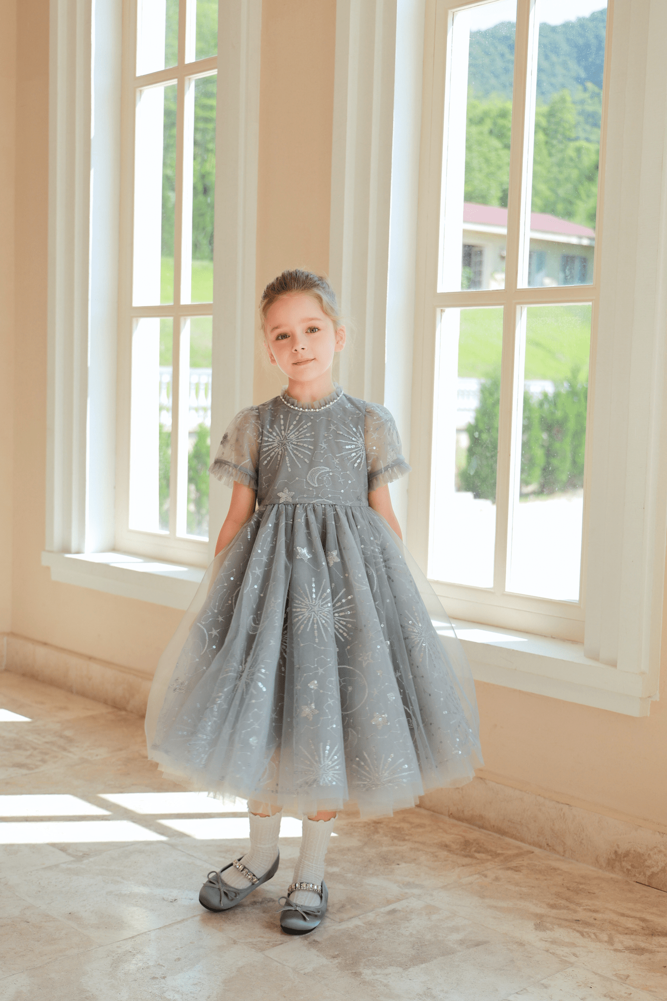 A young girl stands indoors by large windows, sunlight streaming in as she wears the KIKI and JOJO Girls Sequin Embroidered Tulle Dress in Starlight Silver.