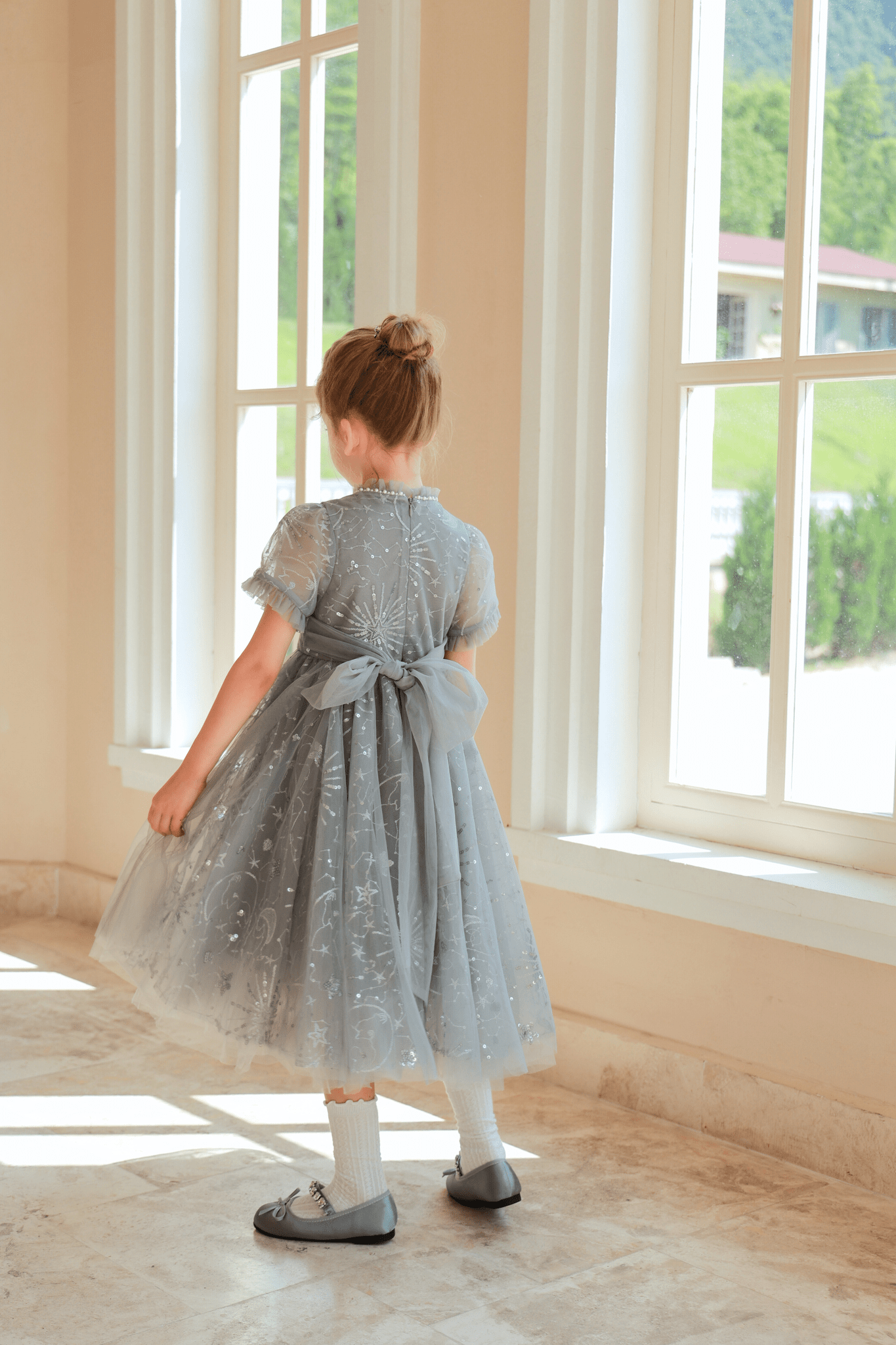 A young girl in the KIKI and JOJO Girls Sequin Embroidered Tulle Dress - Starlight Silver stands indoors by tall windows, facing away from the camera.