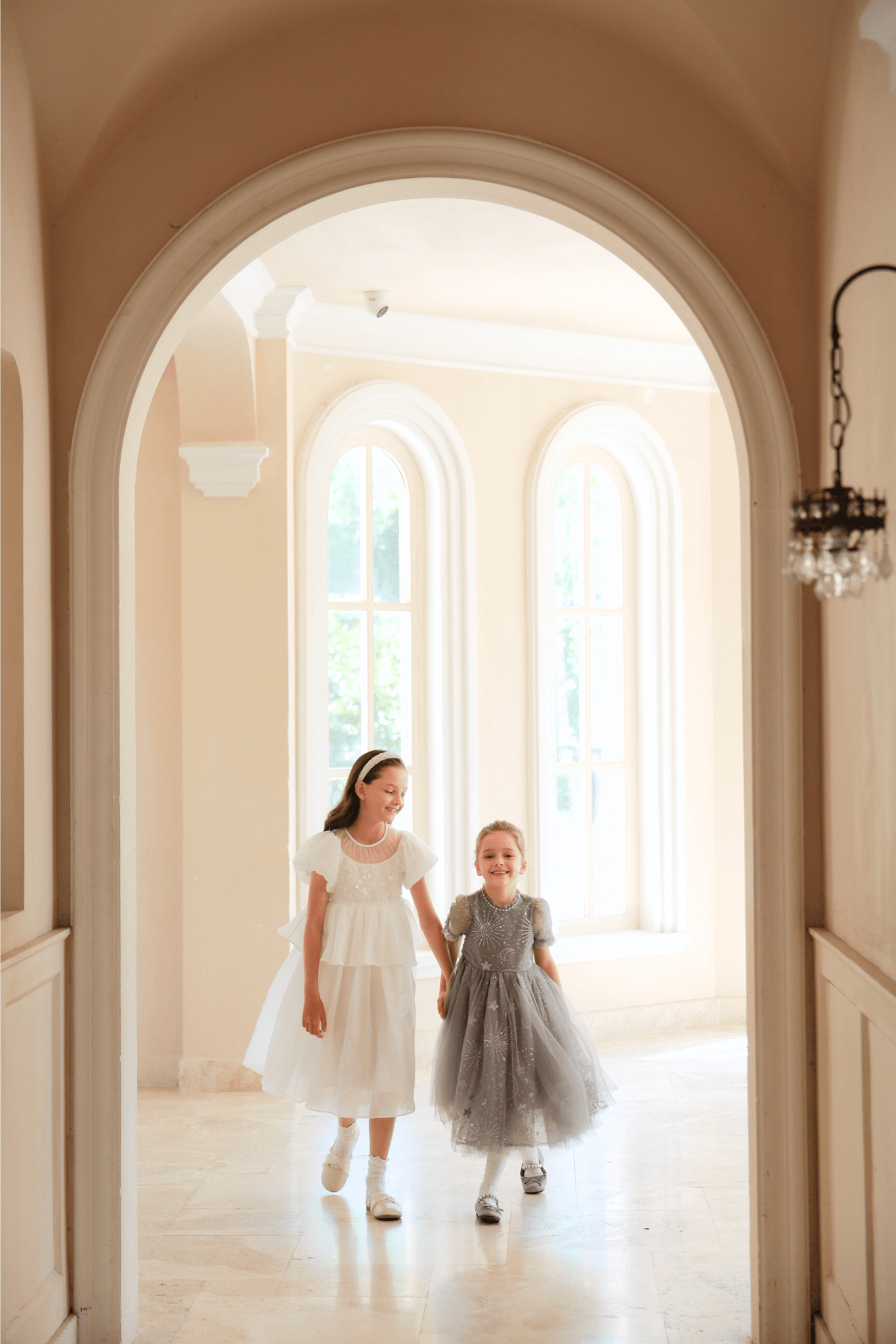 Two young girls in formal dresses, one wearing the KIKI and JOJO Girls Sequin Embroidered Tulle Dress - Starlight Silver, walk hand in hand through an arched hallway with large windows behind them.