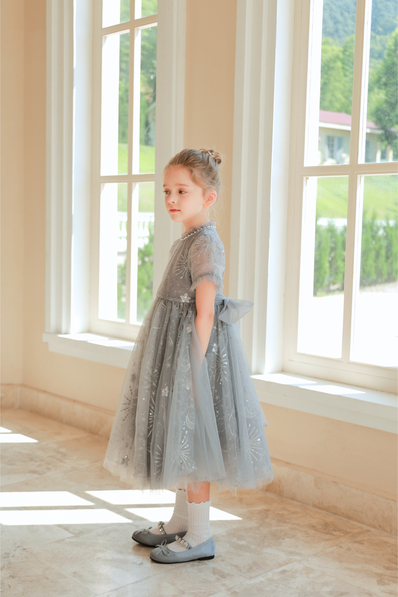 A young girl wears the KIKI and JOJO Girls Sequin Embroidered Tulle Dress in Starlight Silver, standing indoors by large windows with her hands behind her back, looking to the side.