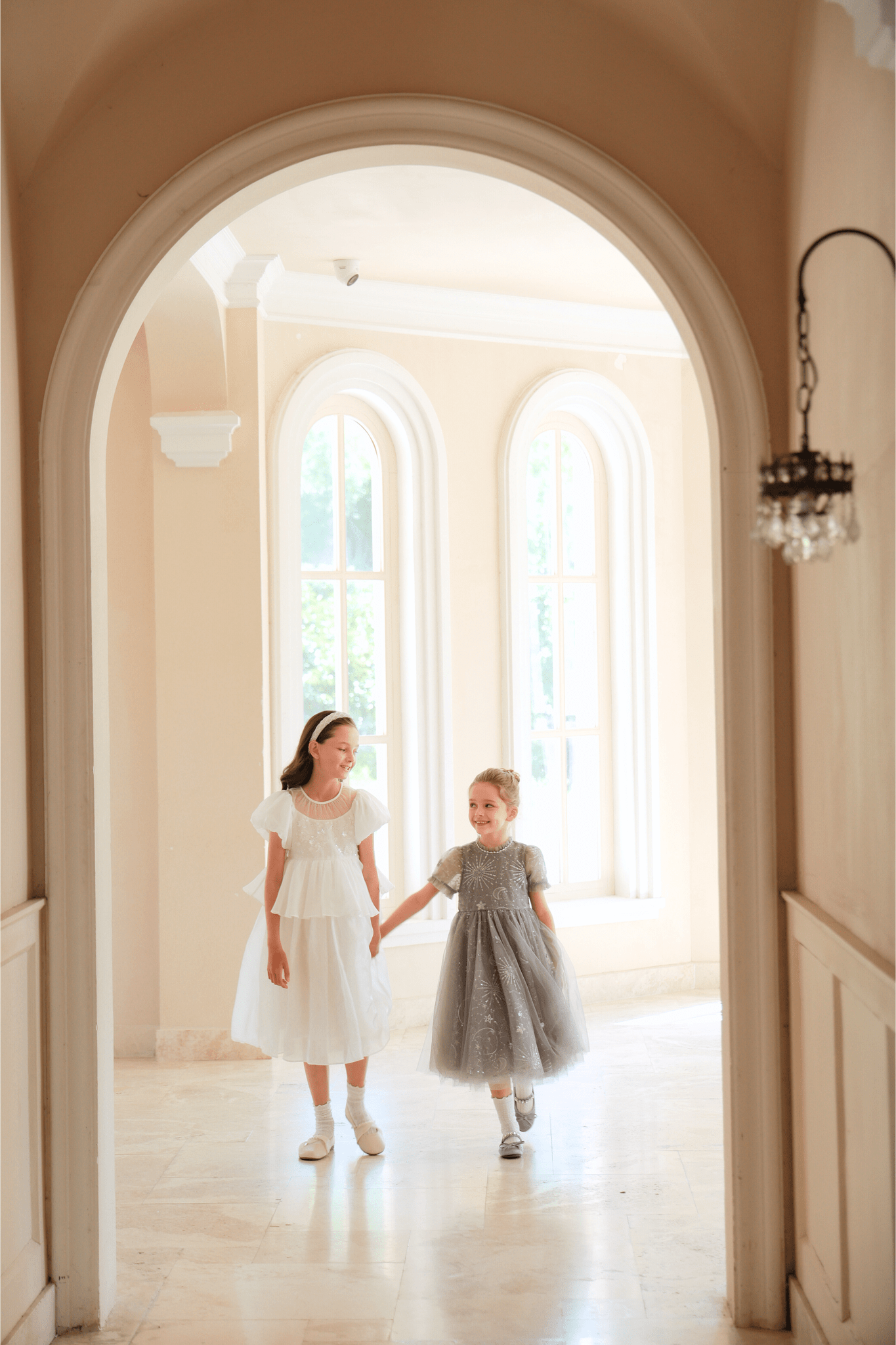 Two young girls walk hand in hand through a bright hallway. One wears the KIKI and JOJO Girls Sequin Embroidered Tulle Dress in Starlight Silver, featuring sparkling sequins and elegant tulle.