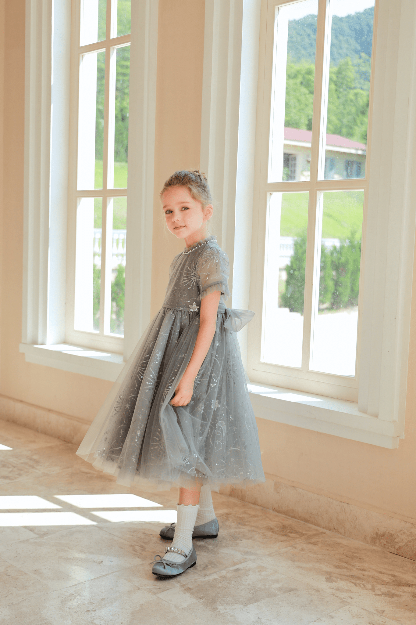A young girl wears the KIKI and JOJO Girls Sequin Embroidered Tulle Dress in Starlight Silver, paired with white socks and grey shoes, standing by large windows with natural light streaming in as she gazes over her shoulder.