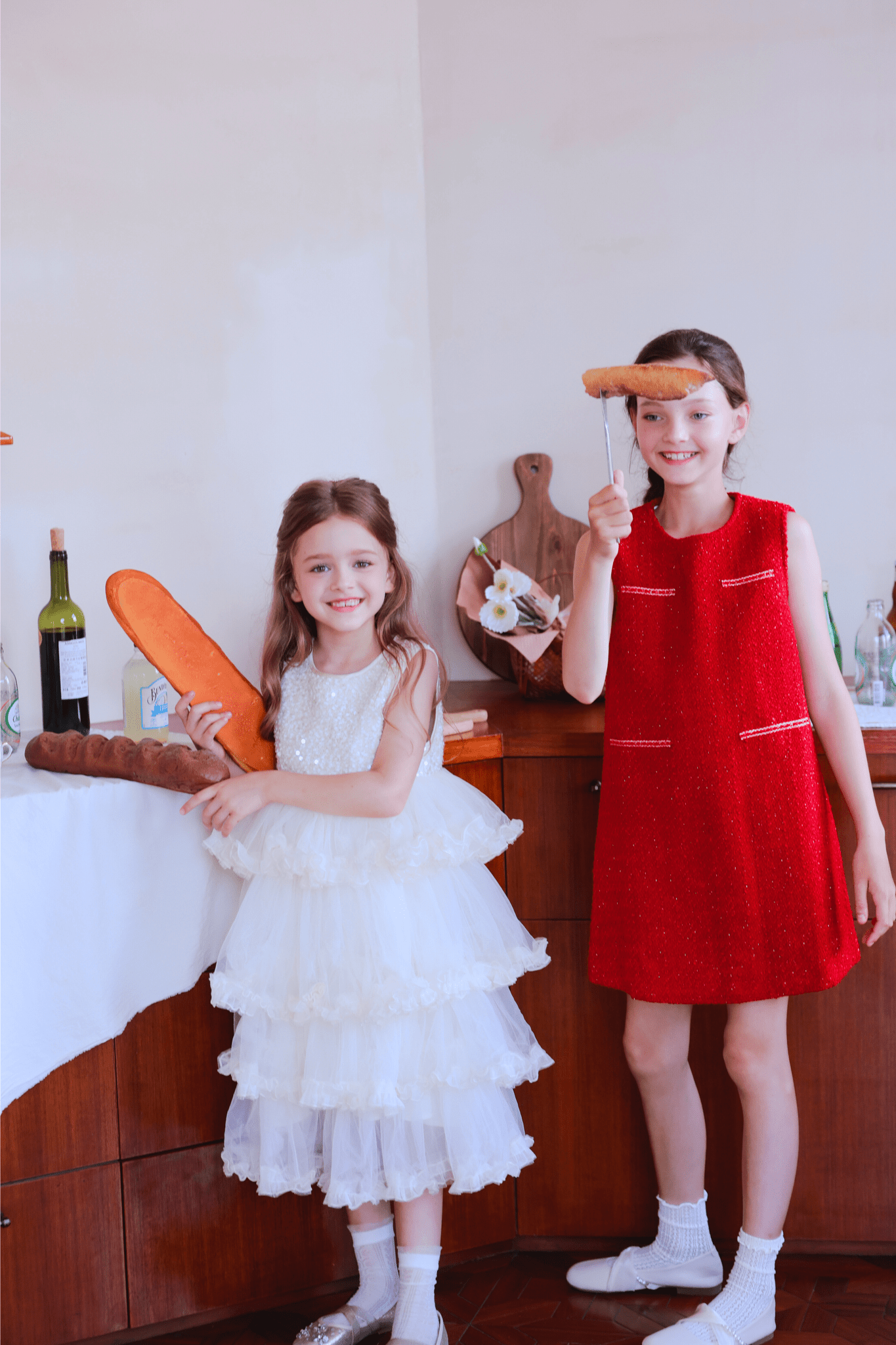 Indoors, two young girls celebrate: one in a white dress holds baguettes, while the other smiles in the KIKI and JOJO Girls Tweed Dress - Cranberry Red, holding bread on a stick; bottles and bread sit on the table behind them.