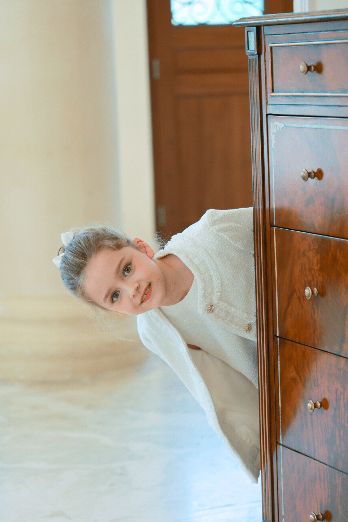 A young girl in a white outfit, wearing a KIKI and JOJO Girls Silk Satin Ribbon Bow Hair Clip in Ivory, peeks out from behind a polished wooden dresser in a bright room with marble floors and a column.