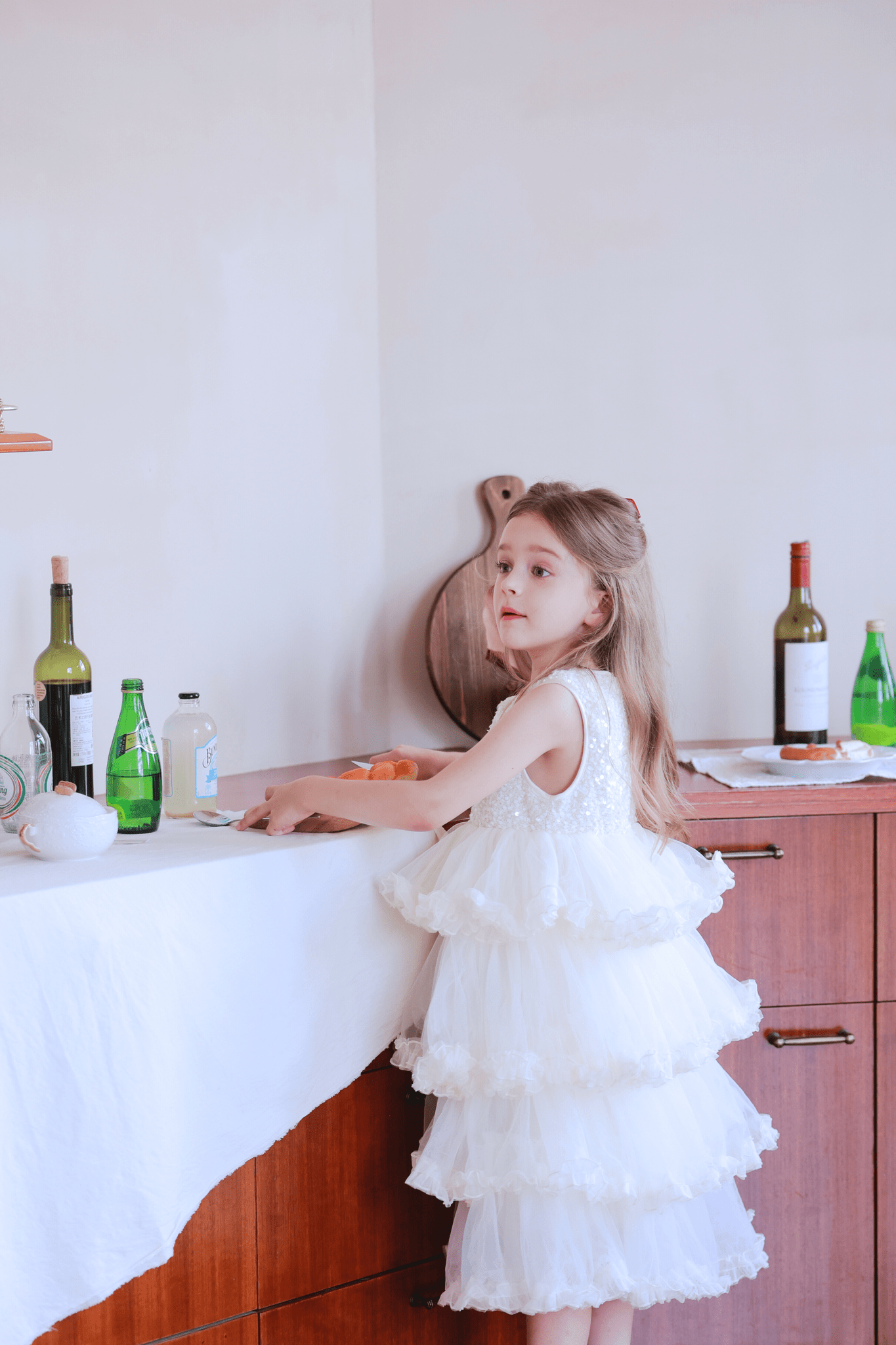 A young girl in a KIKI and JOJO Girls Sparkle Sleeveless Tulle Dress in Champagne stands by a wooden counter adorned with bottles and glassware.
