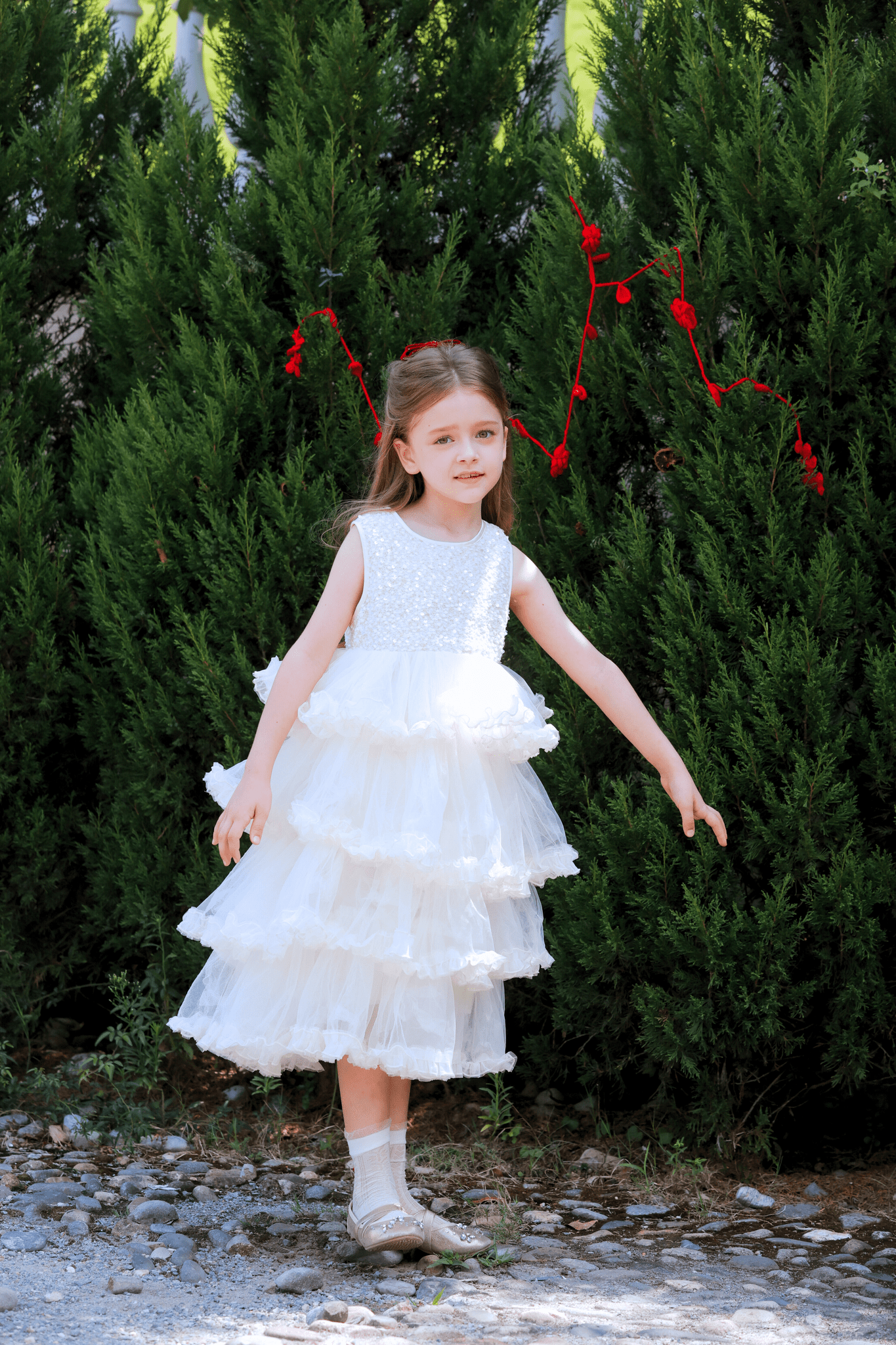 A young girl wears the KIKI and JOJO Girls Sparkle Sleeveless Tulle Dress in Champagne, standing outdoors on a gravel path before tall green bushes, with red string decorations in the background.