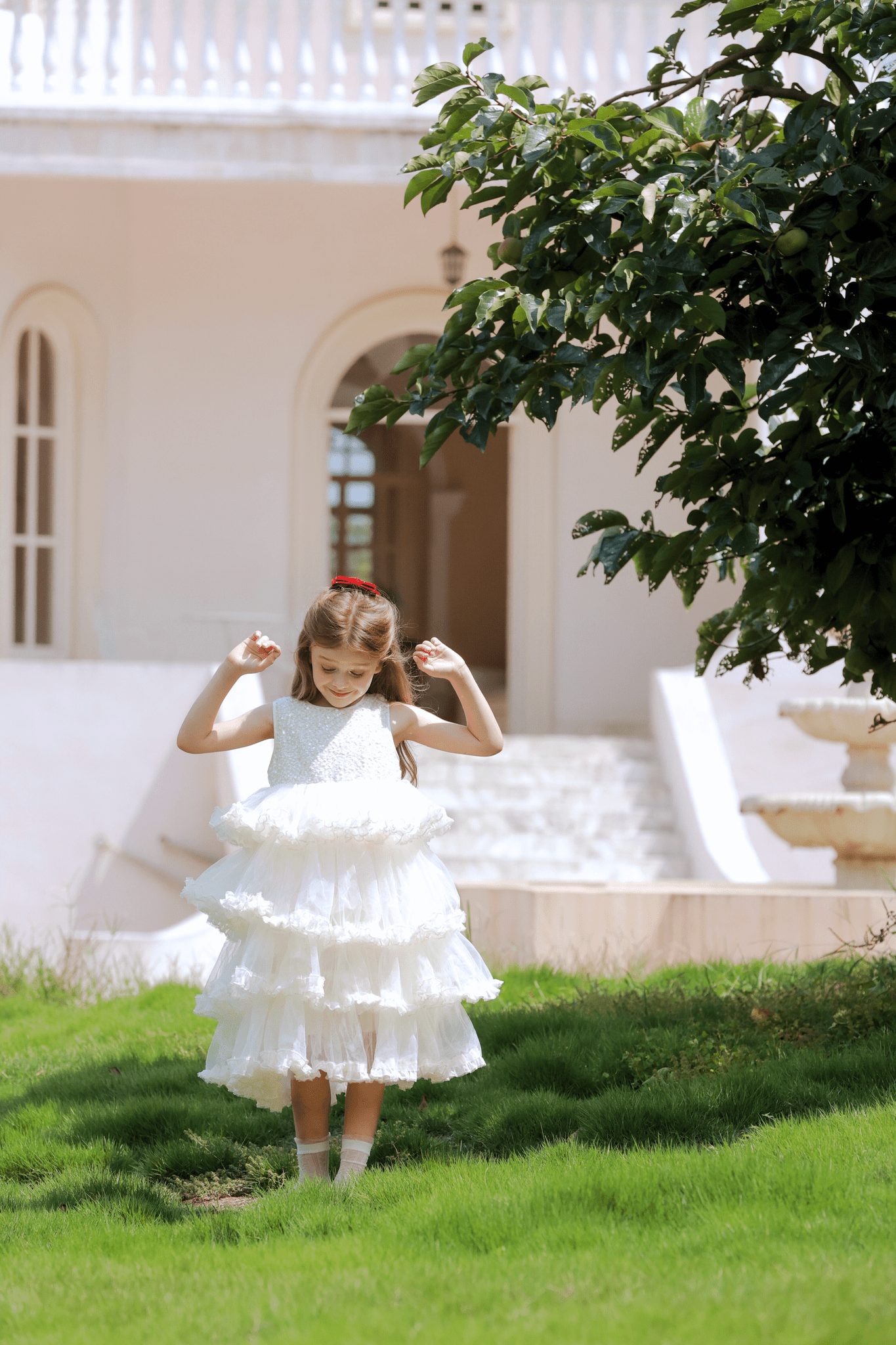 A young girl wears the KIKI and JOJO Girls Sparkle Sleeveless Tulle Dress in champagne as she stands on green grass near a tree, with a white building and fountain visible in the background.