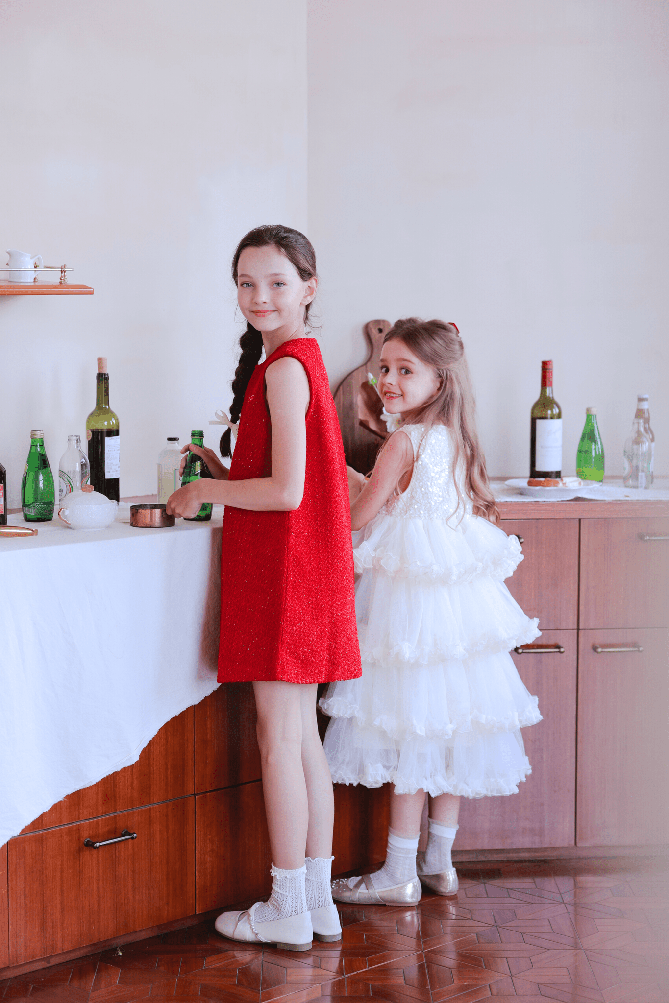 Two young girls stand by a table with bottles and glasses. One wears the KIKI and JOJO Girls Sparkle Sleeveless Tulle Dress in Champagne, while the other is in a white flower girl dress; both look toward the camera.