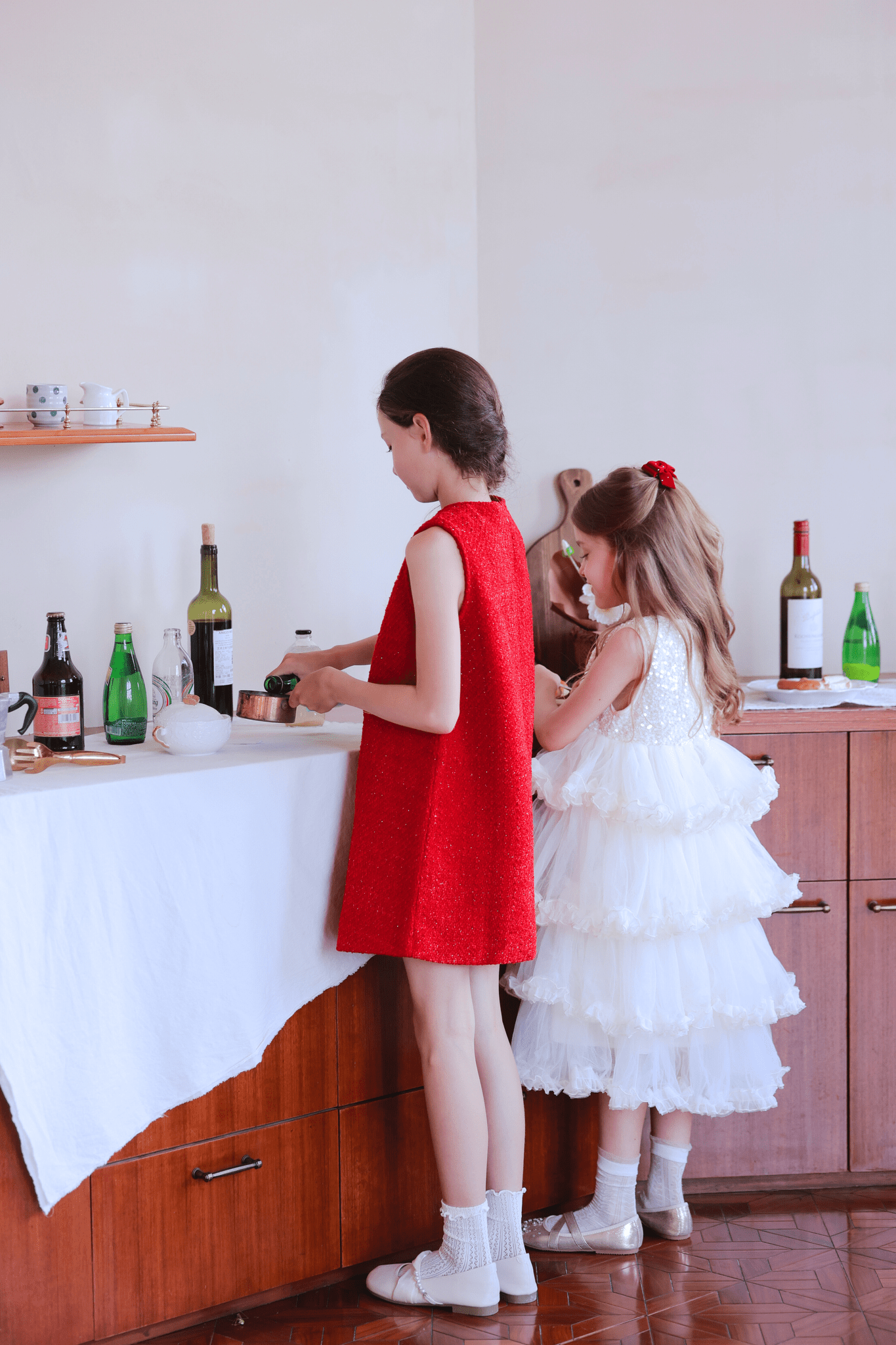 Two young girls in KIKI and JOJO's Girls Sparkle Sleeveless Tulle Dress - Champagne stand at a table with a white cloth, preparing food or drinks among bottles and utensils in a cozy home setting.