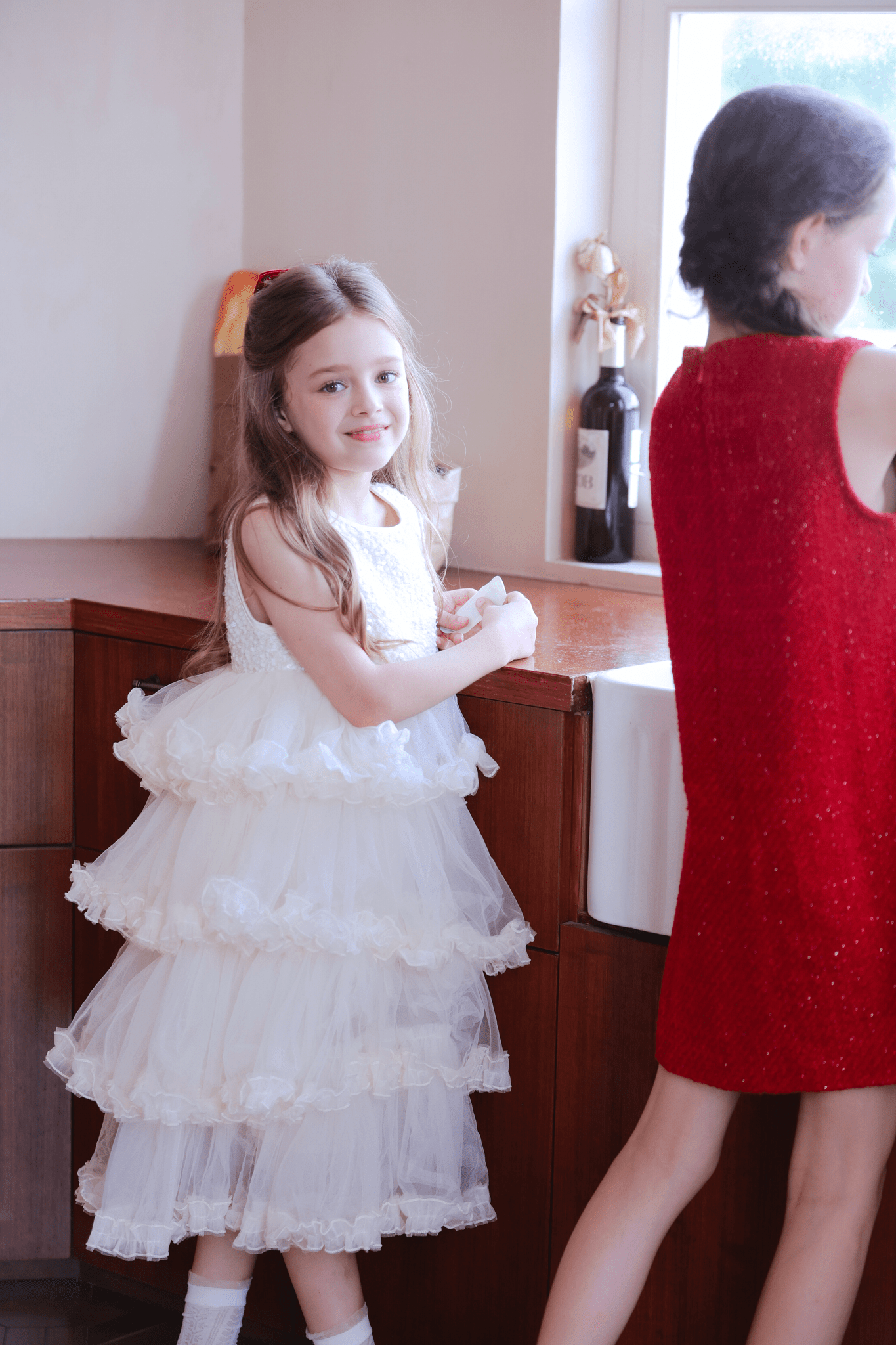 A girl in a KIKI and JOJO Girls Sparkle Sleeveless Tulle Dress - Champagne stands by the kitchen counter, while another girl in a red dress faces away near the sink.