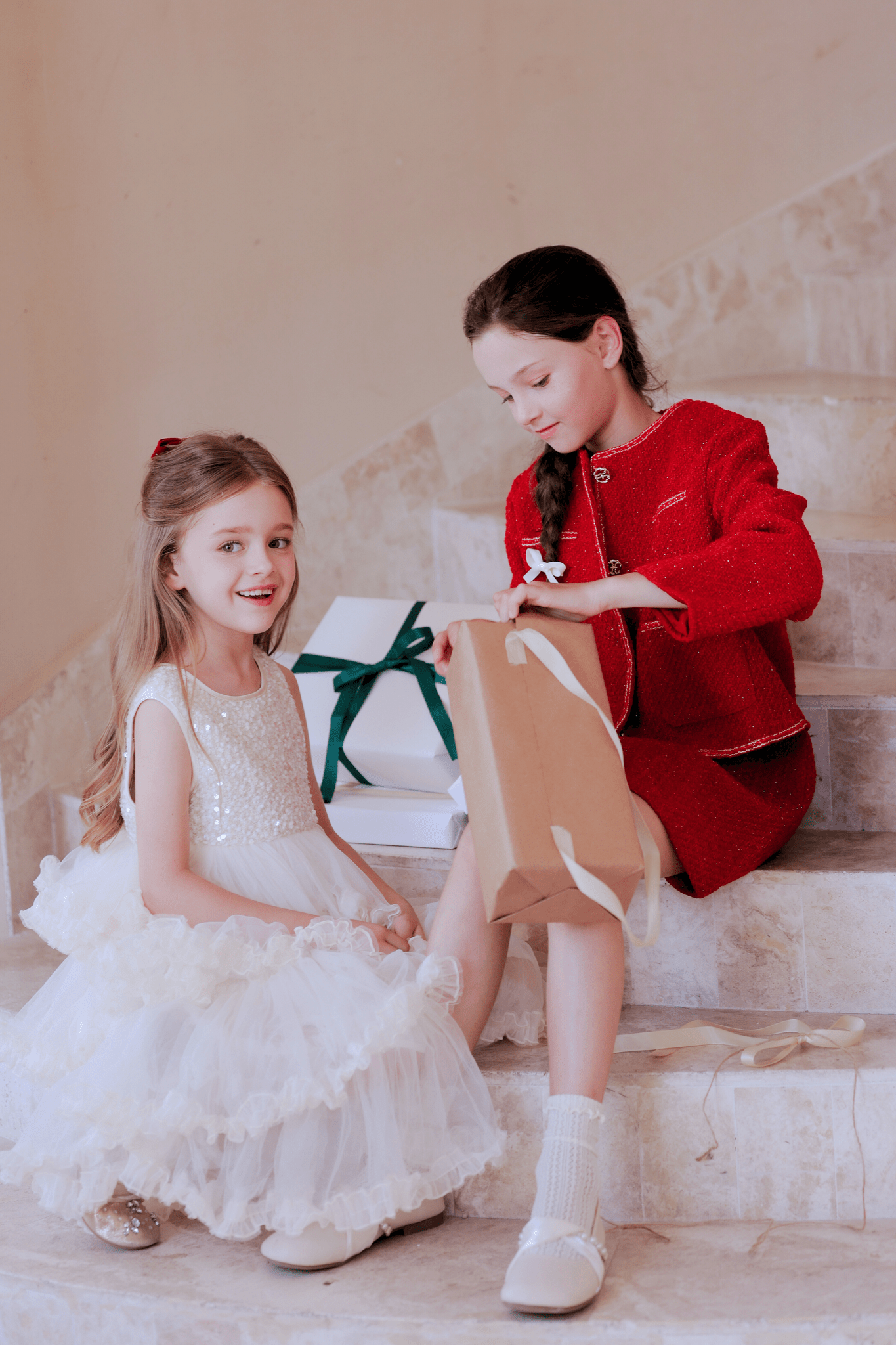 Two young girls sit on marble stairs with gift boxes; one smiles at the camera in a KIKI and JOJO Girls Sparkle Sleeveless Tulle Dress - Champagne, while the other, in a red outfit, opens a brown paper bag.