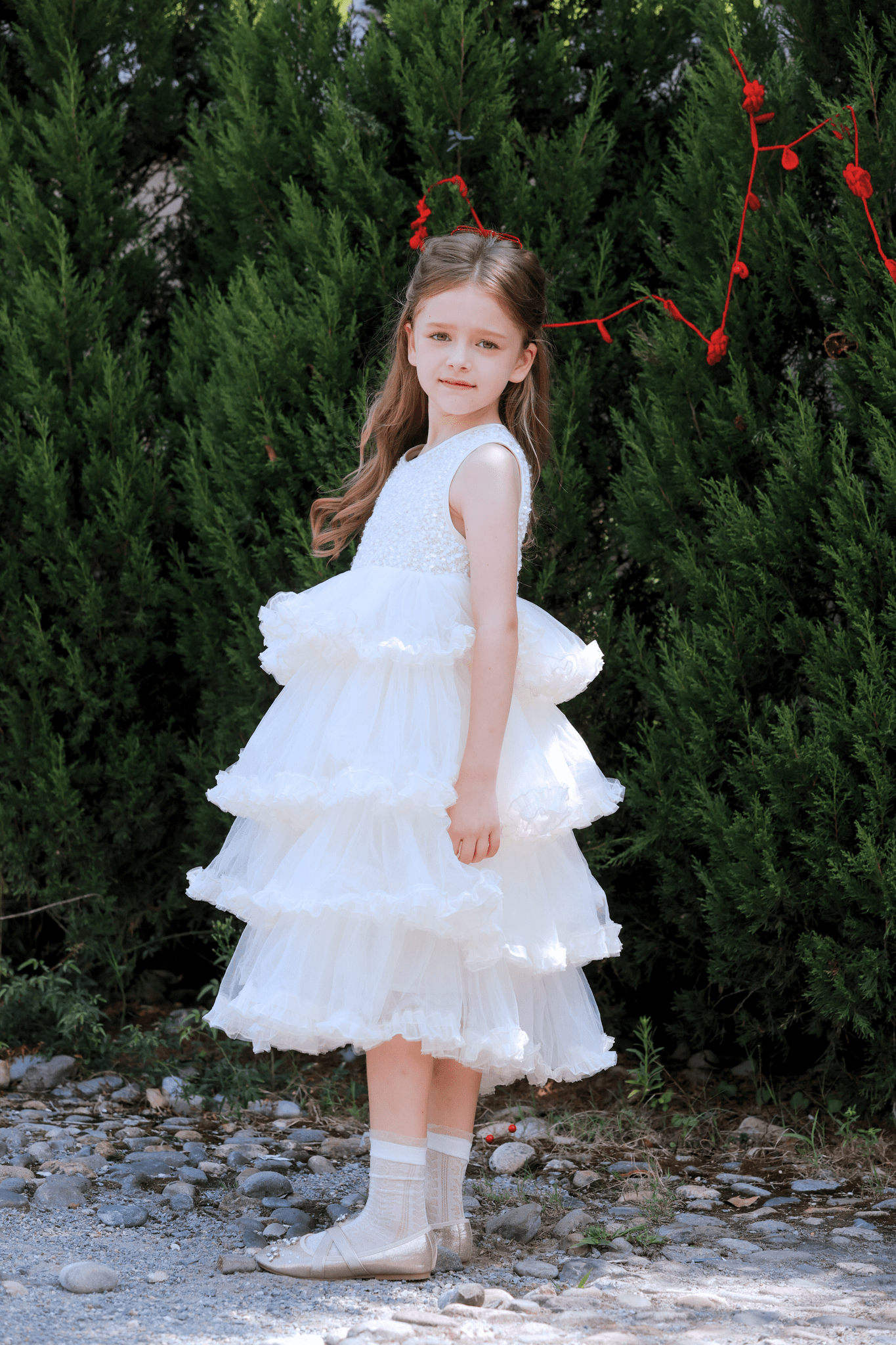 A young girl wears the KIKI and JOJO Girls Sparkle Sleeveless Tulle Dress in Champagne, standing on a gravel path before green bushes and looking at the camera.