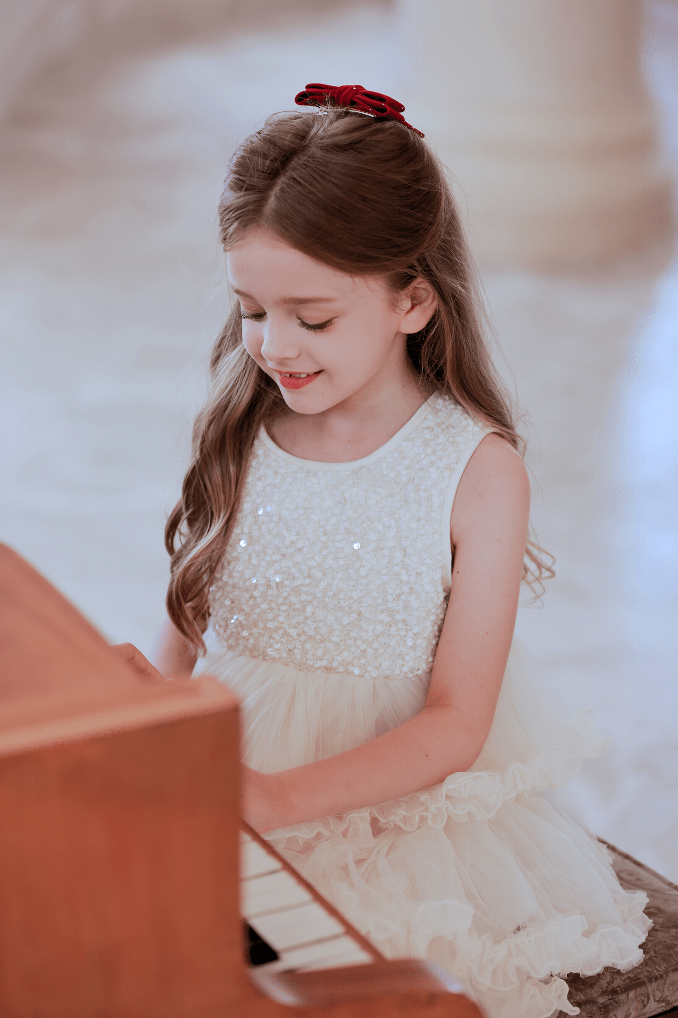 A young girl with long brown hair and a red bow, wearing the KIKI and JOJO Girls Sparkle Sleeveless Tulle Dress in Champagne, sits at a piano and smiles while playing.