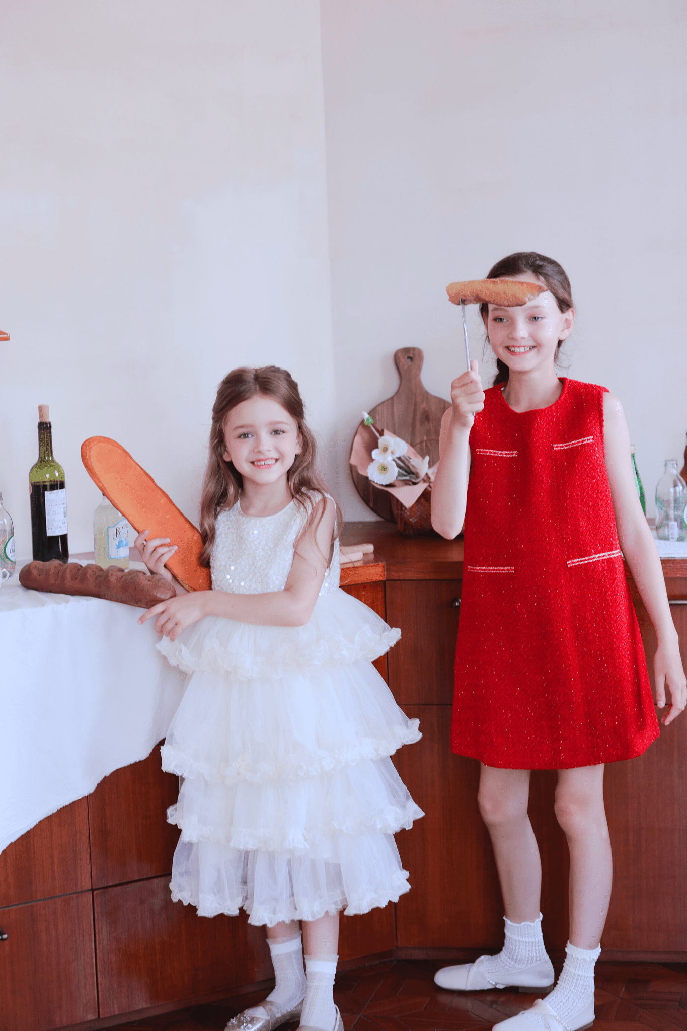 Indoors, one girl in a KIKI and JOJO Girls Sparkle Sleeveless Tulle Dress - Champagne holds bread, while another in a red dress holds a sausage on a fork; bottles and food are on the wooden counter behind them.
