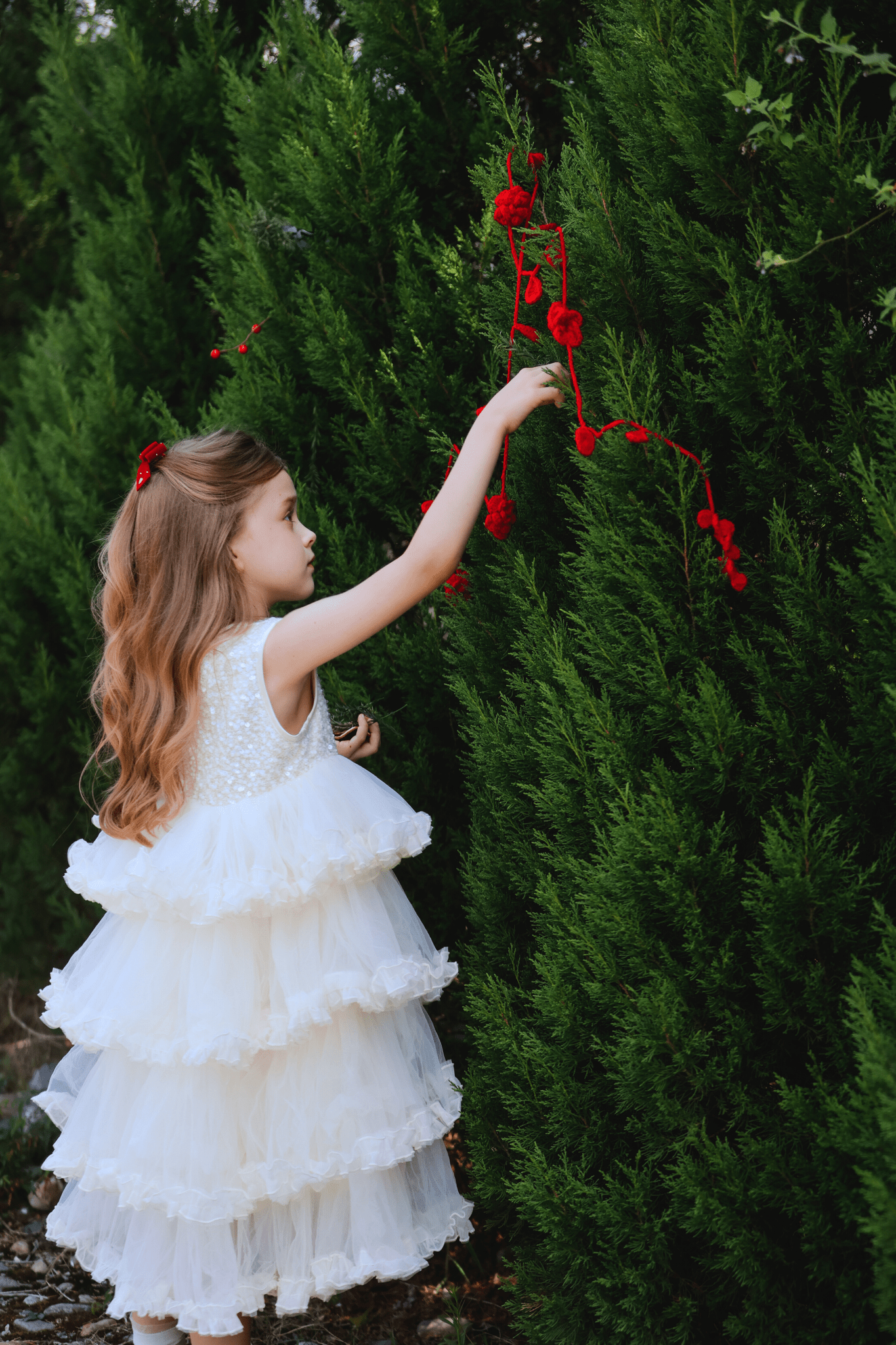 A young girl in a KIKI and JOJO Girls Sparkle Sleeveless Tulle Dress - Champagne hangs red roses on a tall green bush outdoors, her sparkling gown bringing charm to the scene.