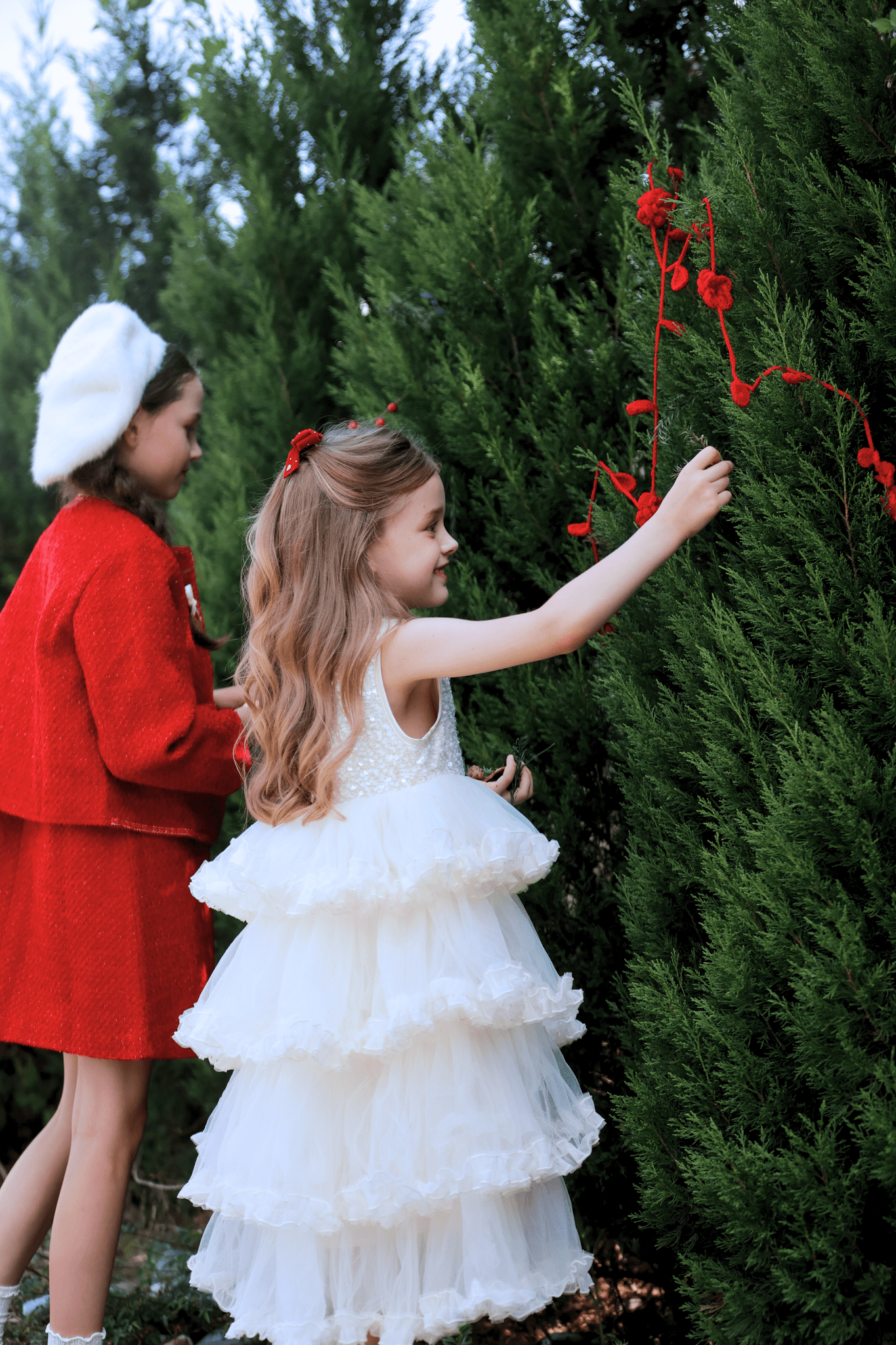 Two young girls, one in a KIKI and JOJO Girls Sparkle Sleeveless Tulle Dress - Champagne and the other in a red coat and beret, decorate an outdoor evergreen bush with red string and ornaments.