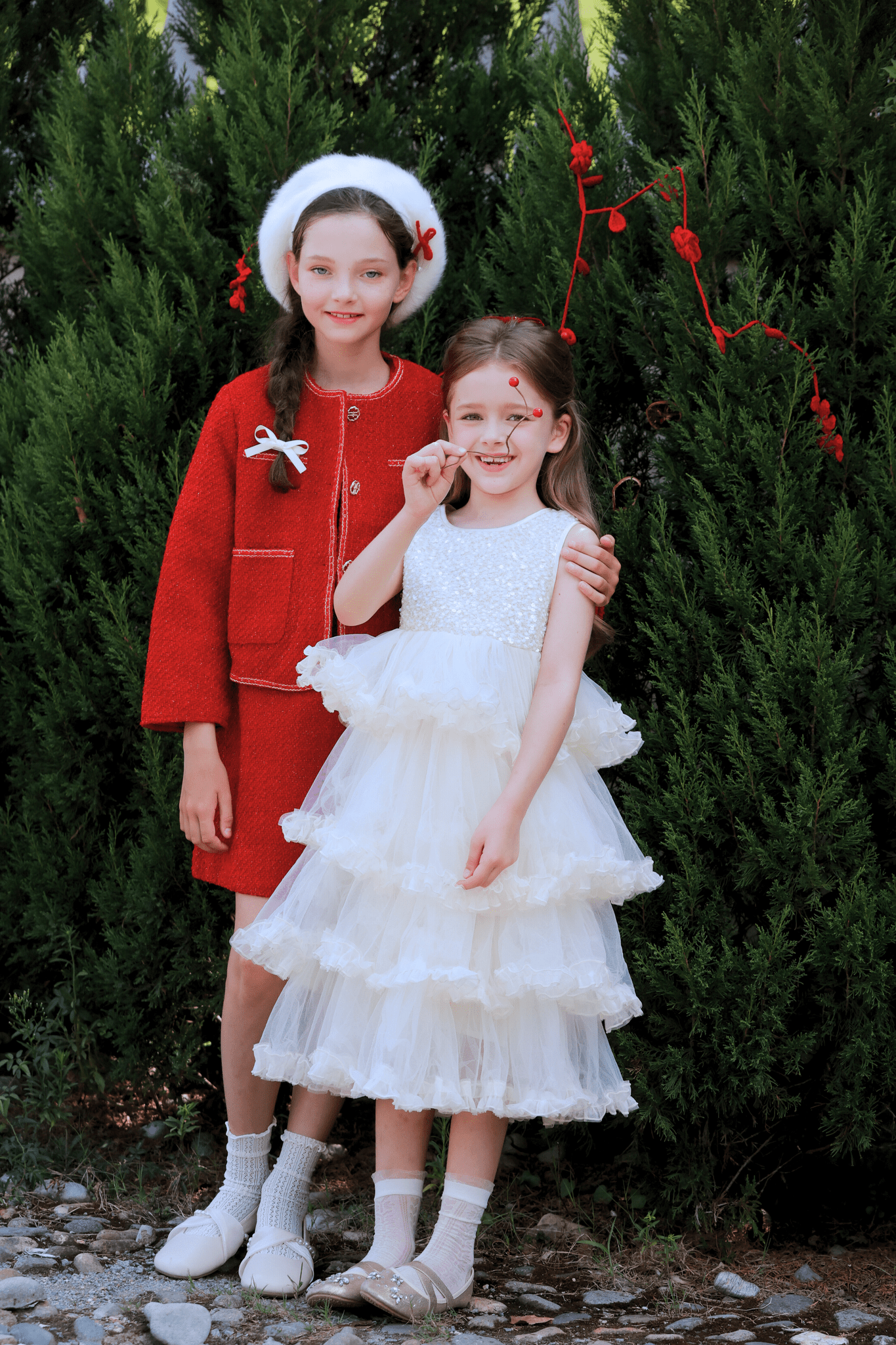 Two young girls pose before green bushes: one in a red coat and white hat, the other beaming in a KIKI and JOJO Girls Sparkle Sleeveless Tulle Dress – Champagne, both smiling brightly at the camera.