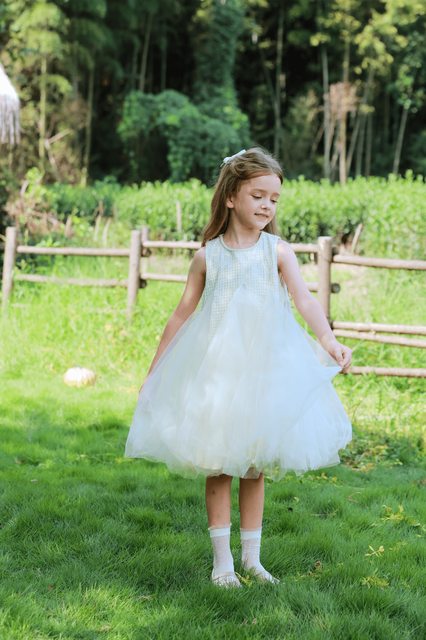 A young girl wears the KIKI and JOJO Girls Textured Velvet Sleeveless Embroidered Dress in Ivory, standing on grass, holding her skirt and smiling with a wooden fence and greenery in the background.