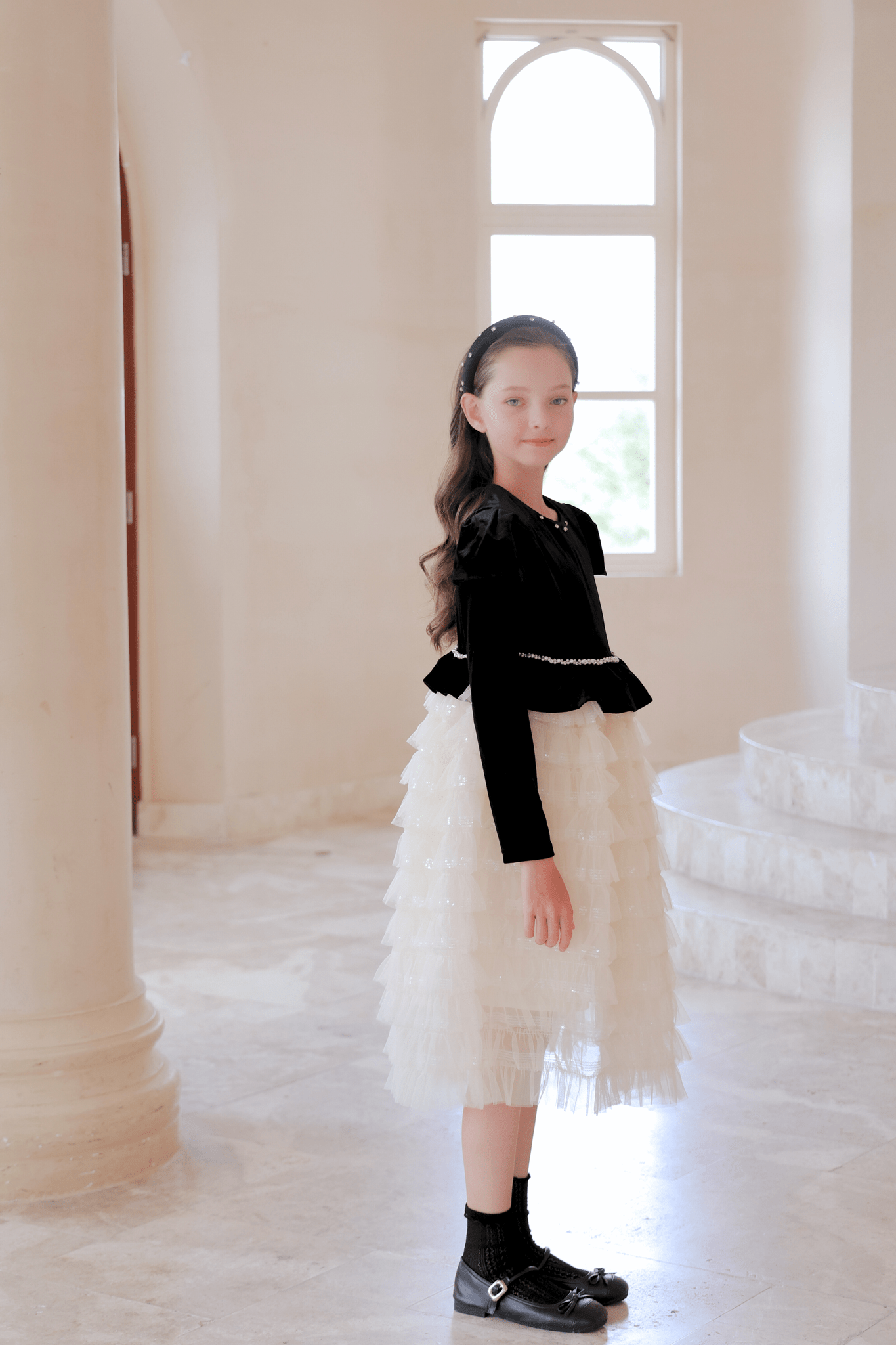 A young girl poses indoors by tall pillars and a window, wearing the KIKI and JOJO Girls Velvet & Tulle Sparkle Dress in Black/Ivory, paired with black shoes and a headband for an elegant, charming look.