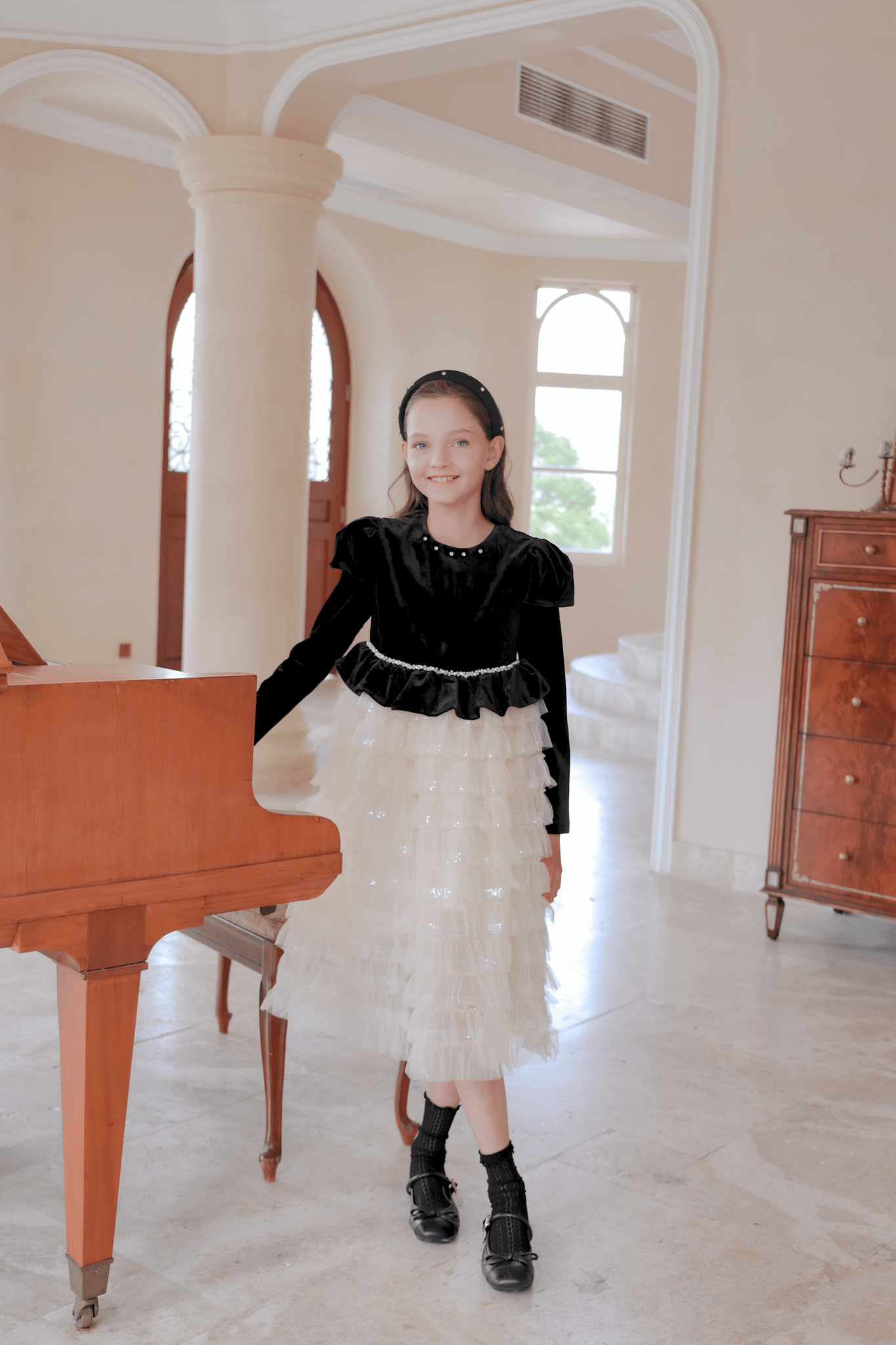 A young girl wearing the KIKI and JOJO Girls Velvet & Tulle Sparkle Dress in Black/Ivory stands beside a wooden piano in a spacious, well-lit room with marble floors.