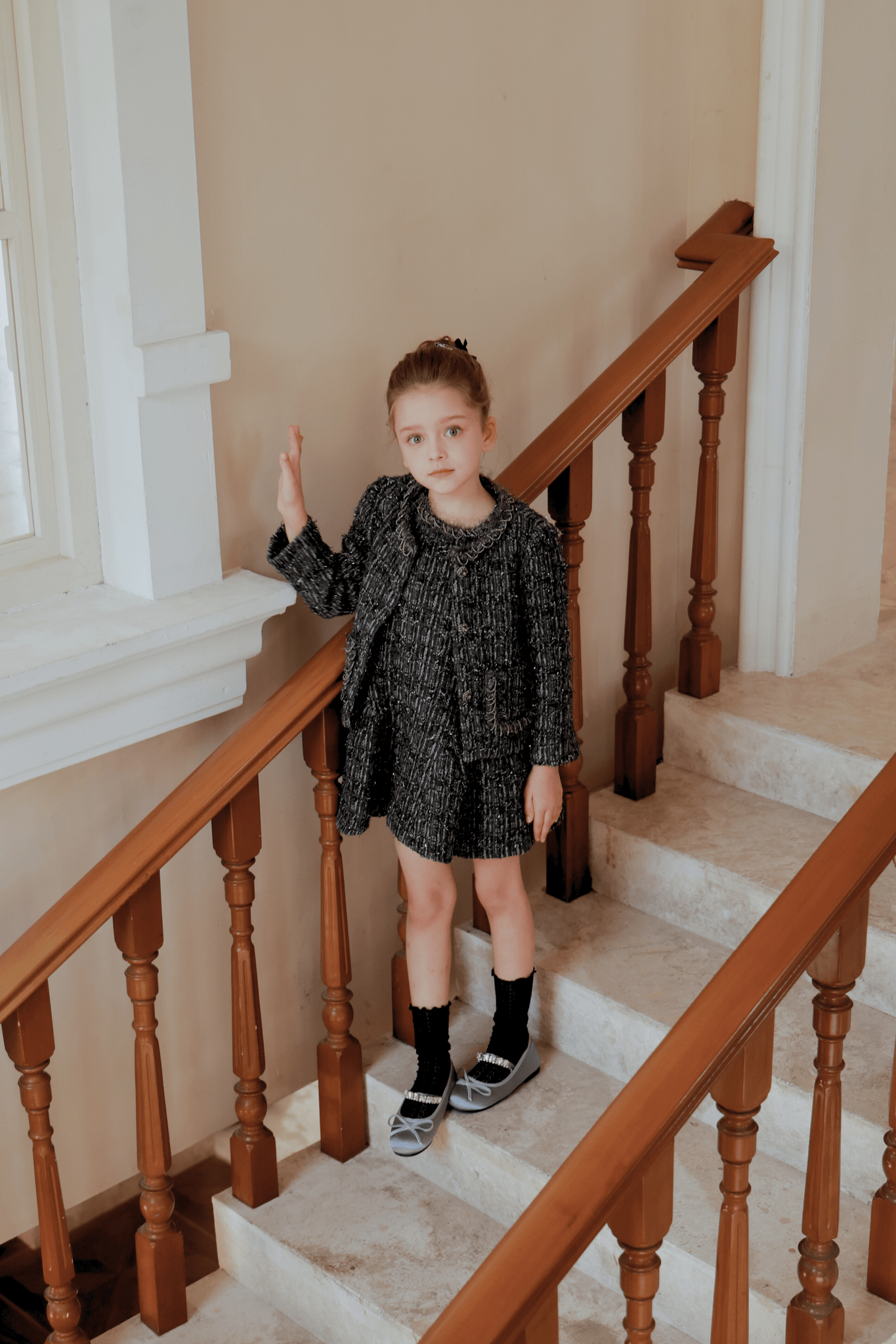 A young girl in the Girls Vintage Beaded Tweed Jacket - Gray by KIKI and JOJO stands on a staircase, hand on railing, looking at the camera and showcasing timeless elegance in sophisticated children's outerwear.