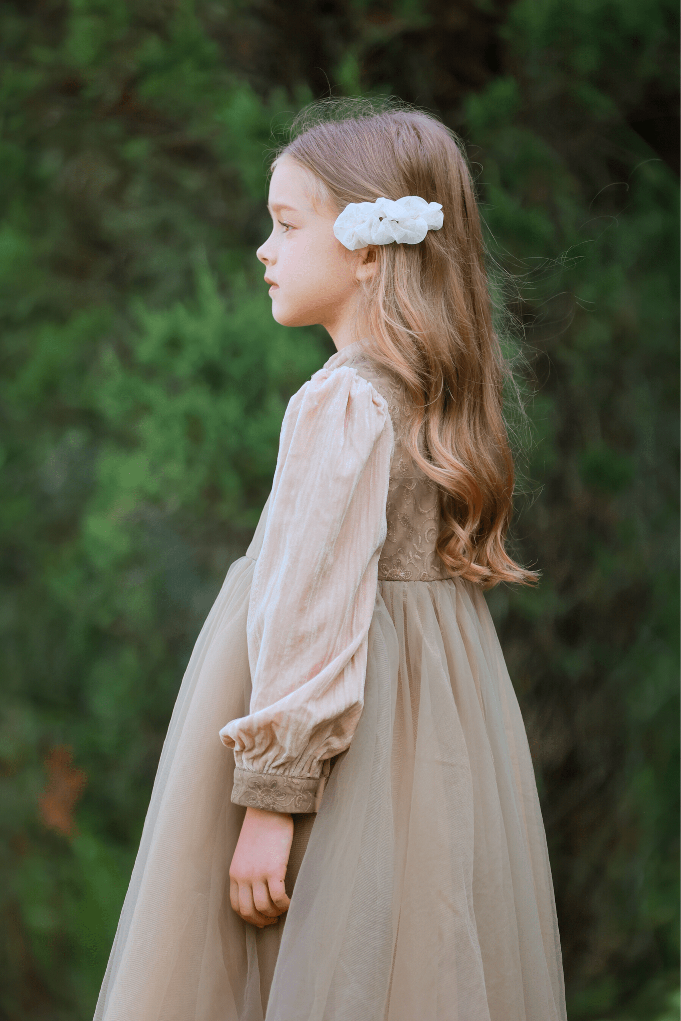 A young girl in profile wears the KIKI and JOJO Girls Vintage Elegance Embroidered Velvet & Tulle Dress, featuring floral embroidery and a white hair accessory, standing outdoors against a green, leafy background—ideal for formal occasions.