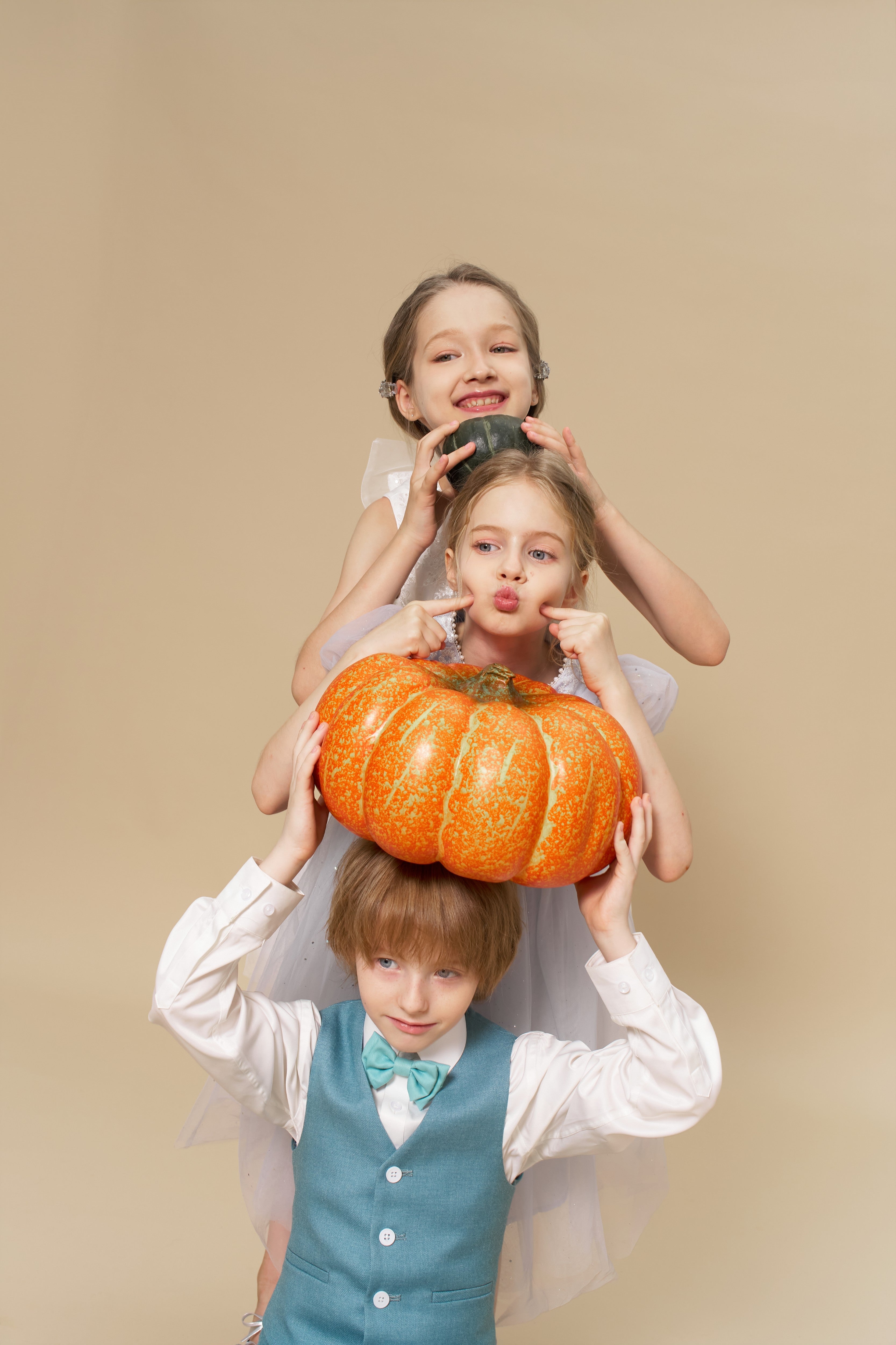 Three children pose playfully in a studio; the boy at the bottom holds a large pumpkin on his head while the two girls above make silly faces.