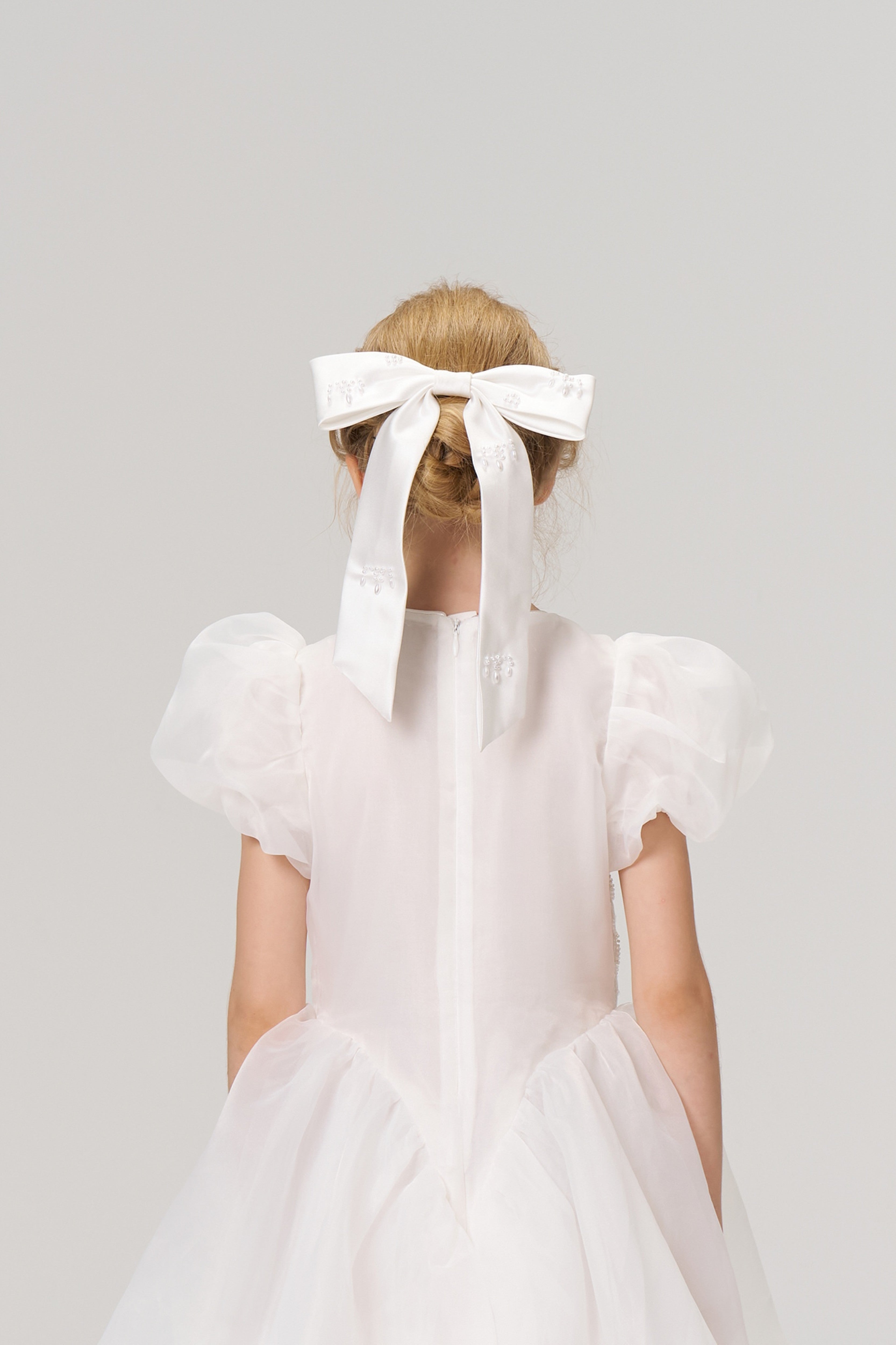 A young girl in a white dress with puffed sleeves stands facing away, wearing a large white bow in her hair.