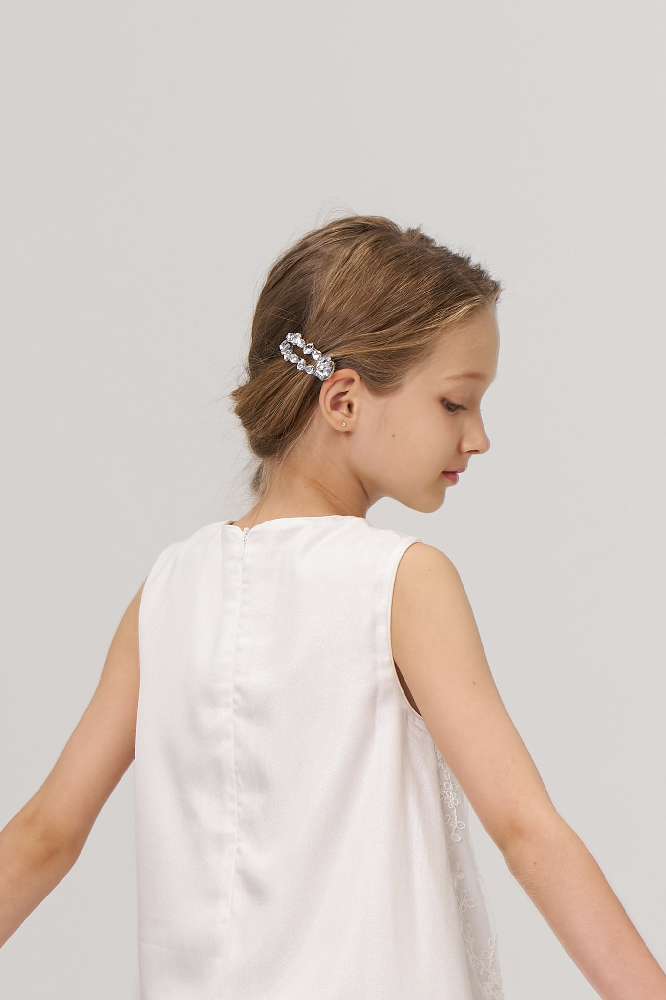 Young girl in a sleeveless white dress with her hair styled back and a decorative hair clip, facing sideways against a plain background.