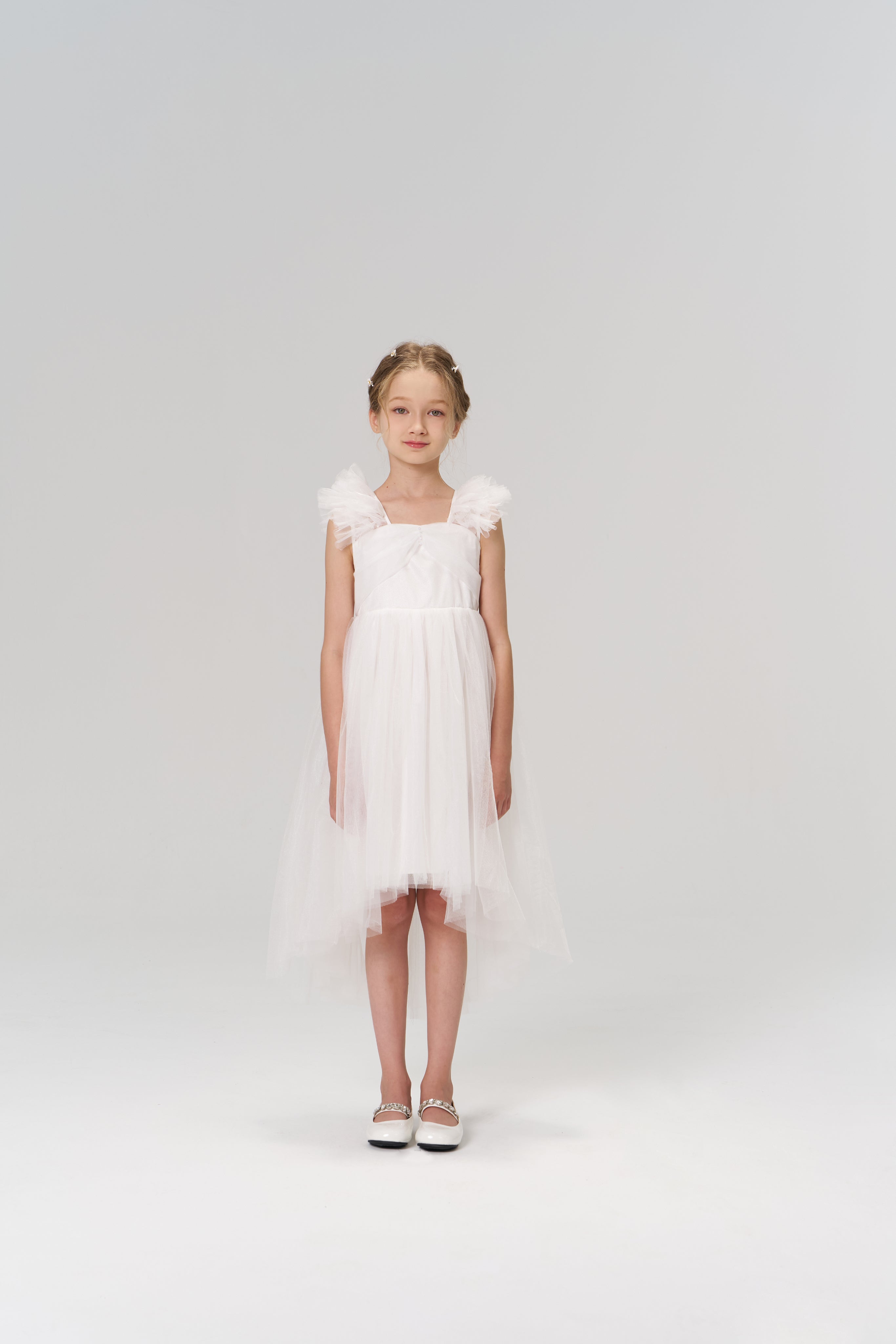 A young girl faces forward wearing the Cloud Whisper Dress by KIKI and JOJO, a flowy white tulle dress with delicate details and white shoes, set against a plain light background.