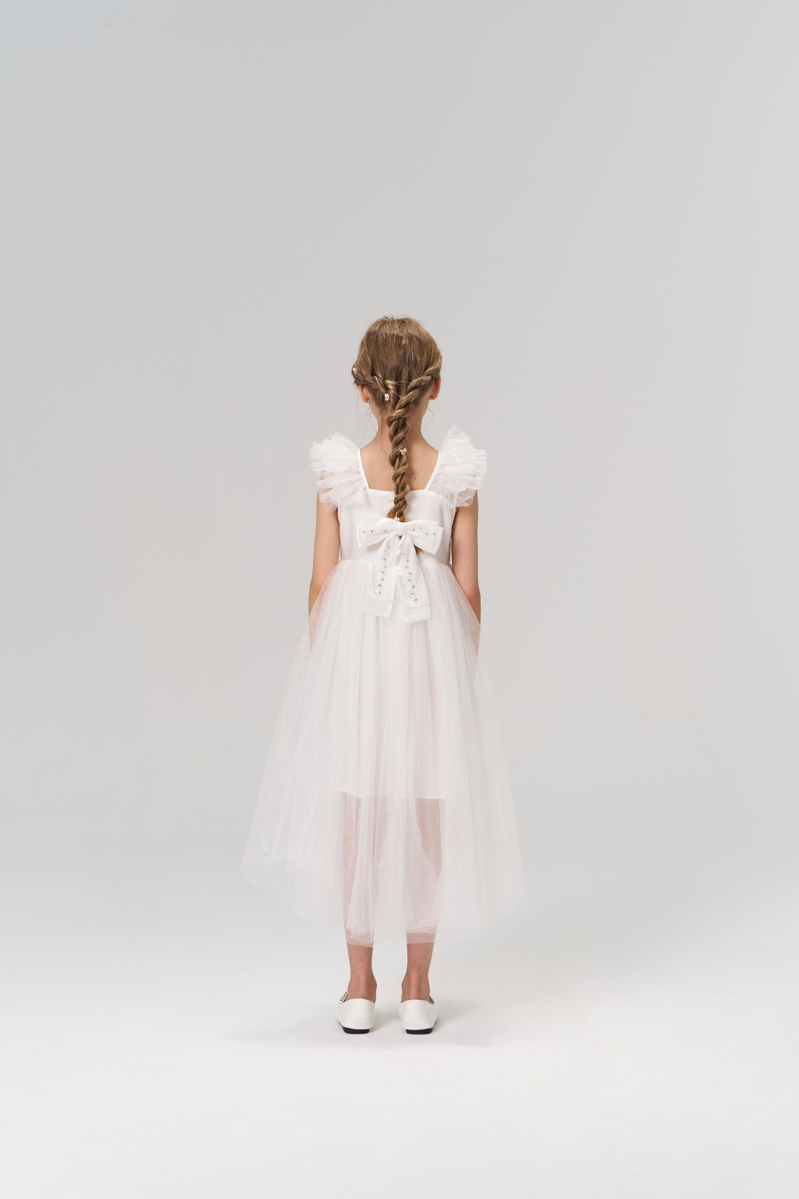 A young girl faces away from the camera, wearing the KIKI and JOJO Cloud Whisper Dress—a flowy white tulle design with a large back bow—set against a plain light background.