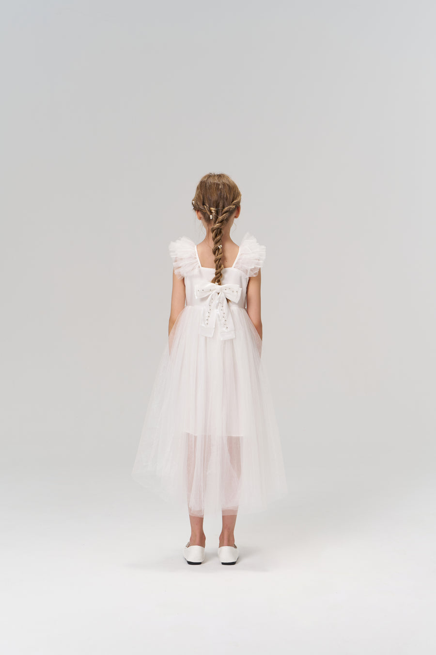 A young girl faces away from the camera, wearing the KIKI and JOJO Cloud Whisper Dress—a flowy white tulle design with a large back bow—set against a plain light background.