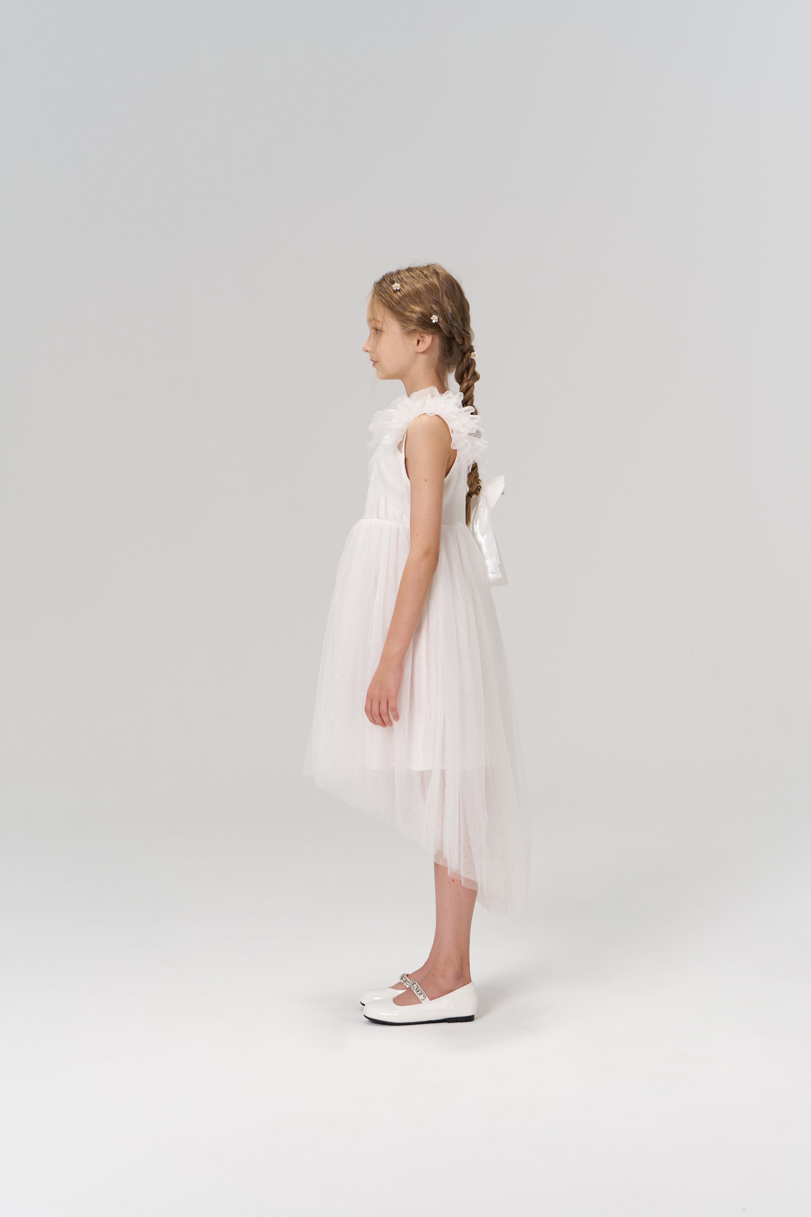 A young girl stands in profile against a plain light background, wearing the Cloud Whisper Dress by KIKI and JOJO, a flowy white tulle dress paired with matching white shoes.