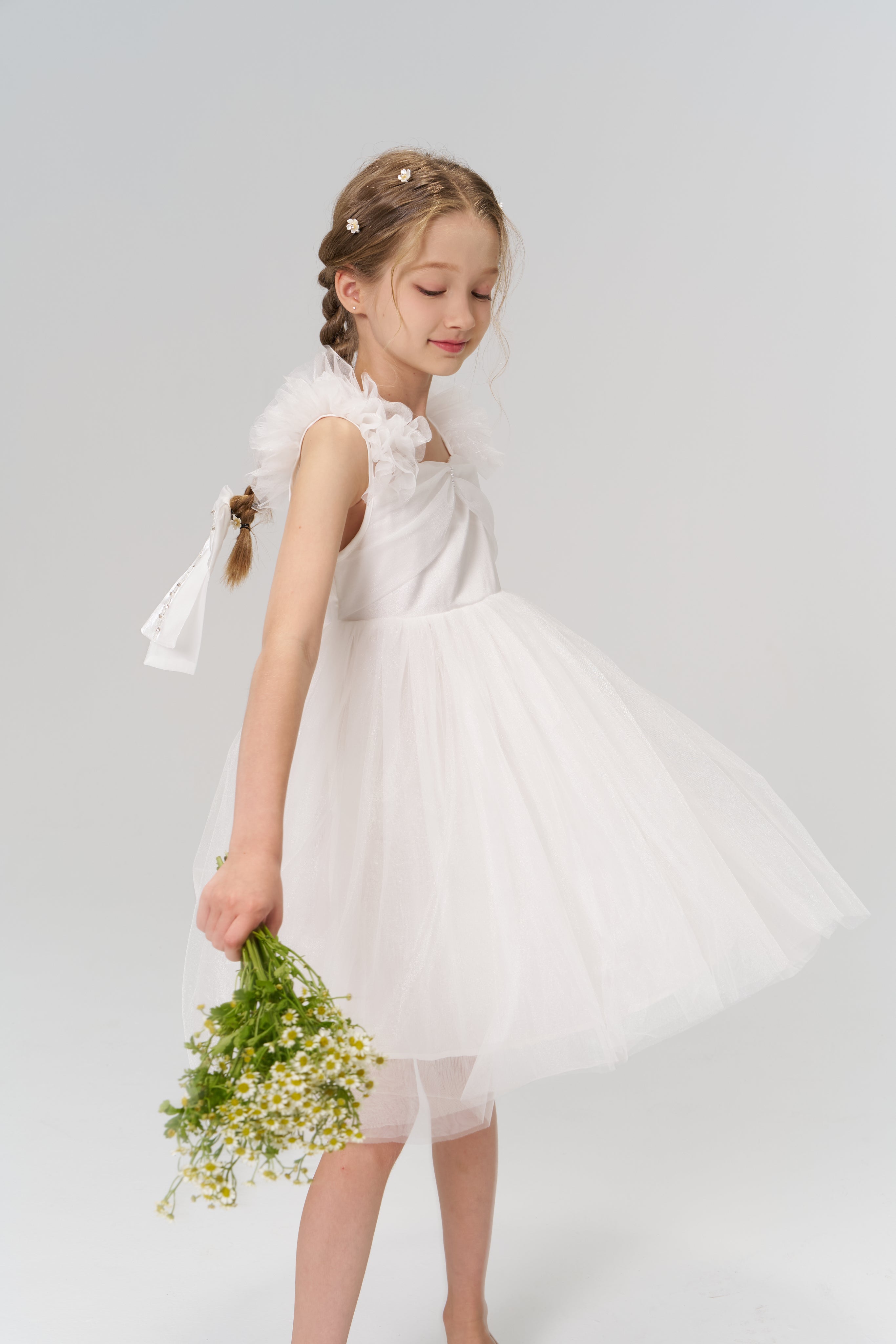 A young girl in a white dress holds a small bouquet of flowers, standing against a plain light background.